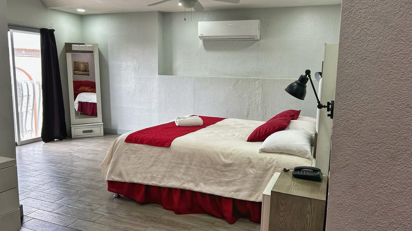 A hotel room with a bed, a desk with a telephone, a mirror, and an air conditioning unit on the wall.