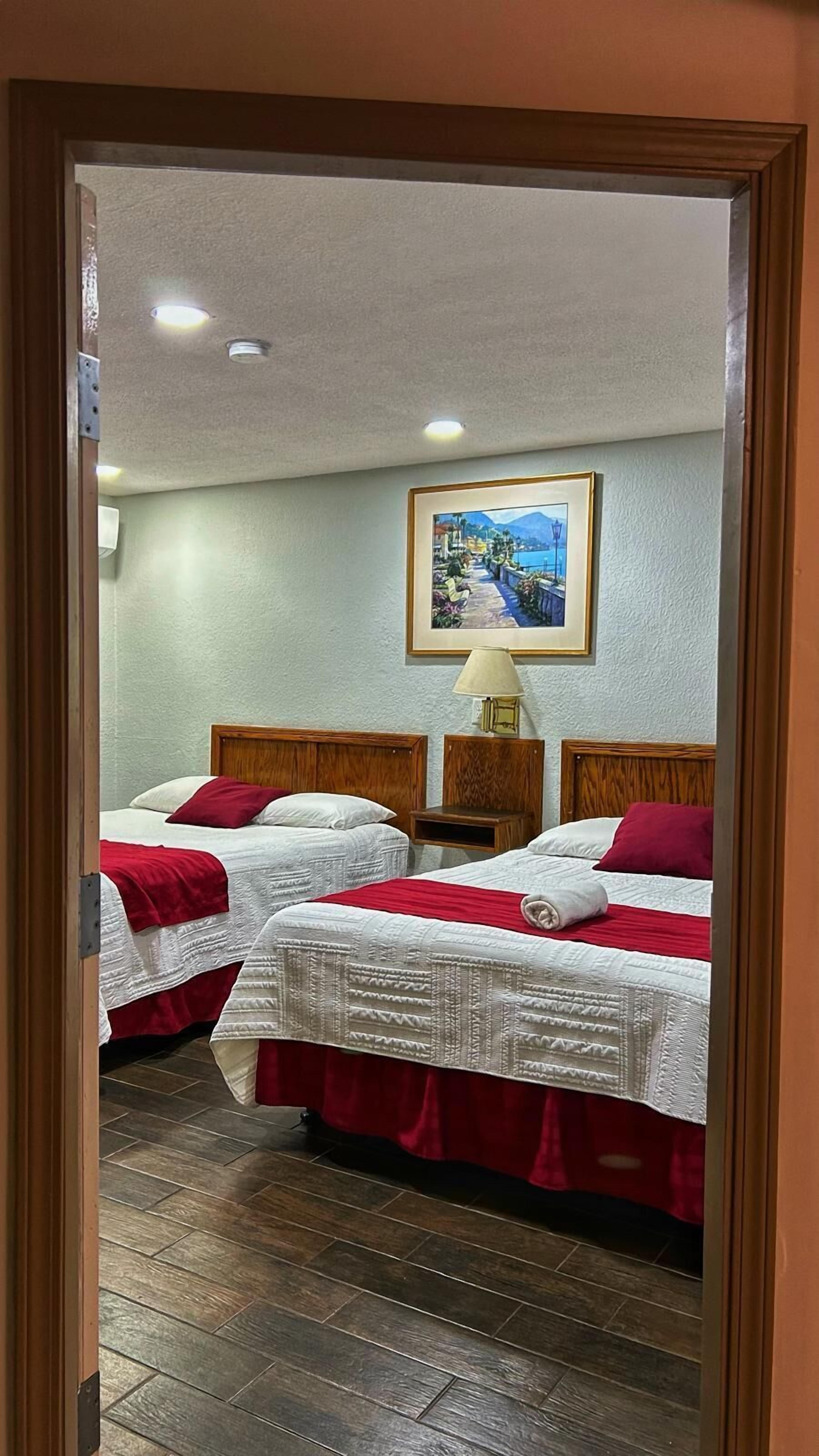 A hotel room with two beds, each with a red and white bedspread, a wooden headboard, and a painting of a coastal scene on the wall.