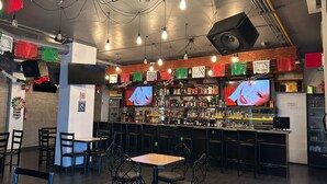 Un espace bar avec un comptoir, des étagères remplies de bouteilles, des luminaires suspendus et un écran de télévision.