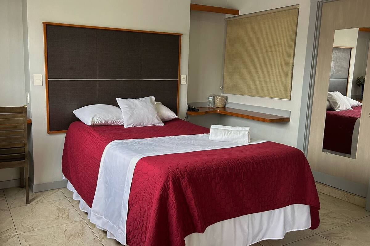A hotel room with a bed, a chair, a small table, a mirror, and a potted plant.