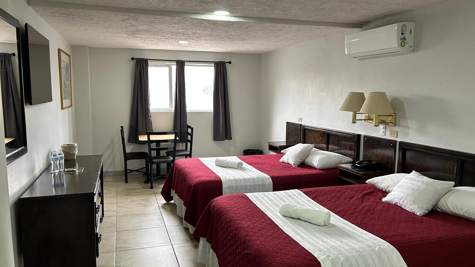 Habitación de hotel con dos camas, un escritorio, una silla, una mesita y un ventanal con cortinas.