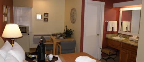 A hotel room with a bed, a desk, a dining table, a microwave, a lamp, a bathroom with a sink and mirror, and towels on the bed.