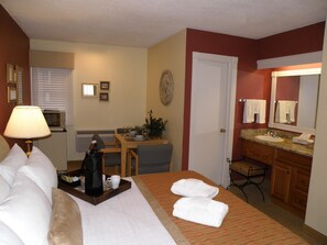A hotel room with a bed, a desk, a dining table, a microwave, a lamp, a bathroom with a sink and mirror, and towels on the bed.