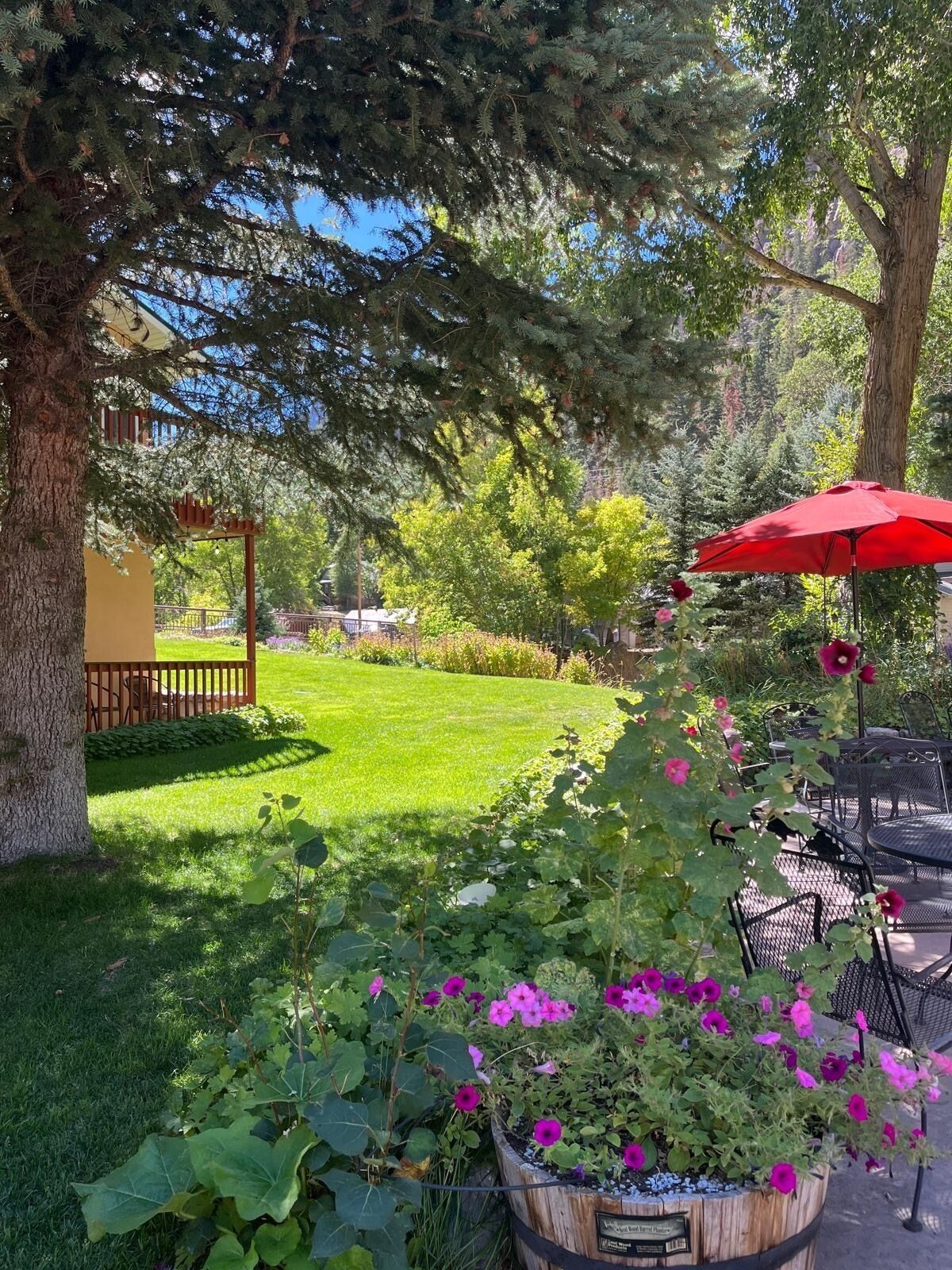 A patio area with a wooden table and chairs, a red umbrella, and a flower bed with pink flowers.
