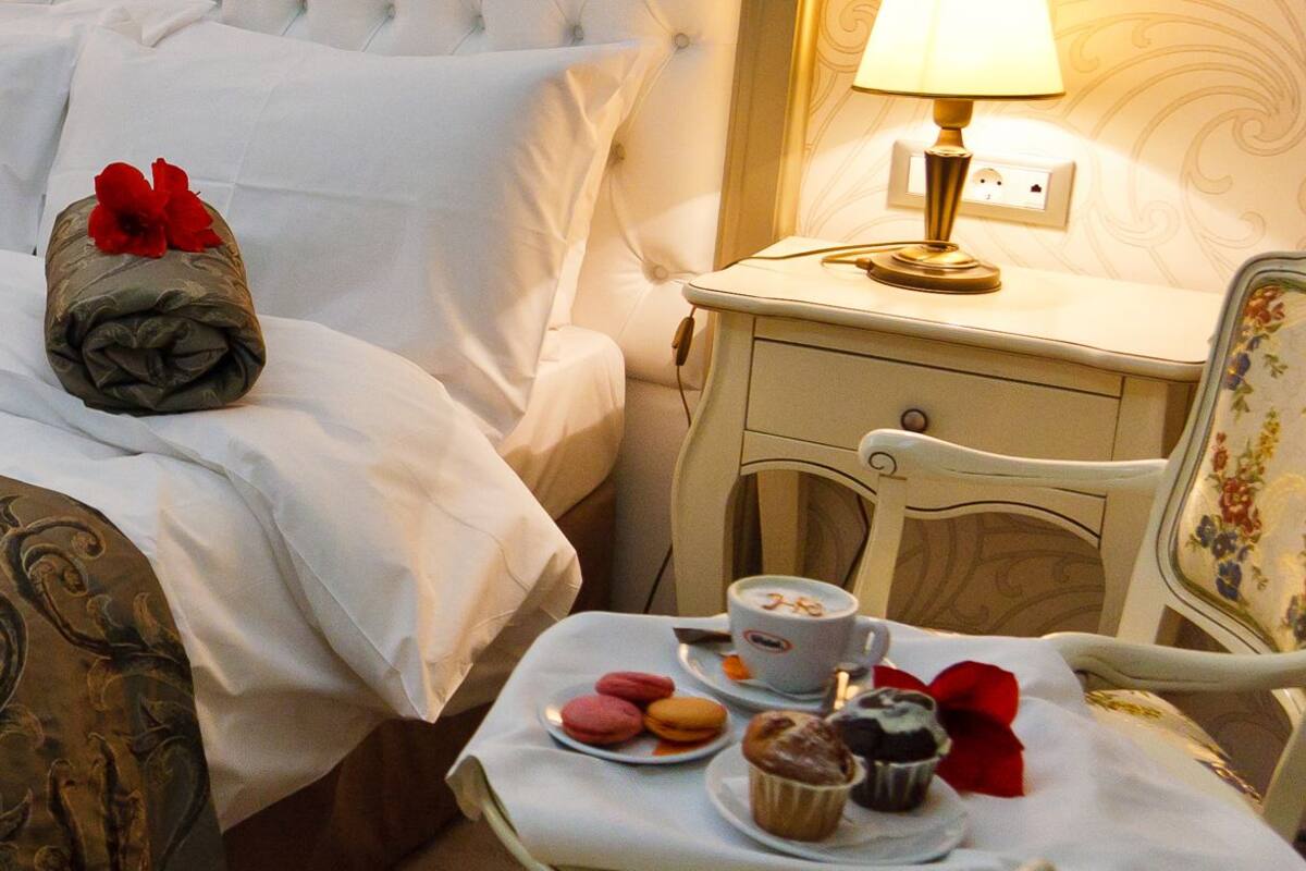 Un lit bien fait, recouvert d’une couvre-lit à motifs, deux serviettes enroulées ornées de fleurs rouges, une table de chevet avec une lampe, une chaise avec un coussin à fleurs et une petite table avec des pâtisseries et une tasse.