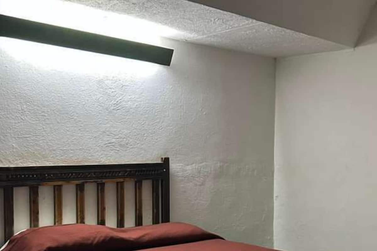 Ein kleines Zimmer mit einem Einzelbett, einem Holzkopfteil, einer roten Bettdecke und einer Deckenleuchte.