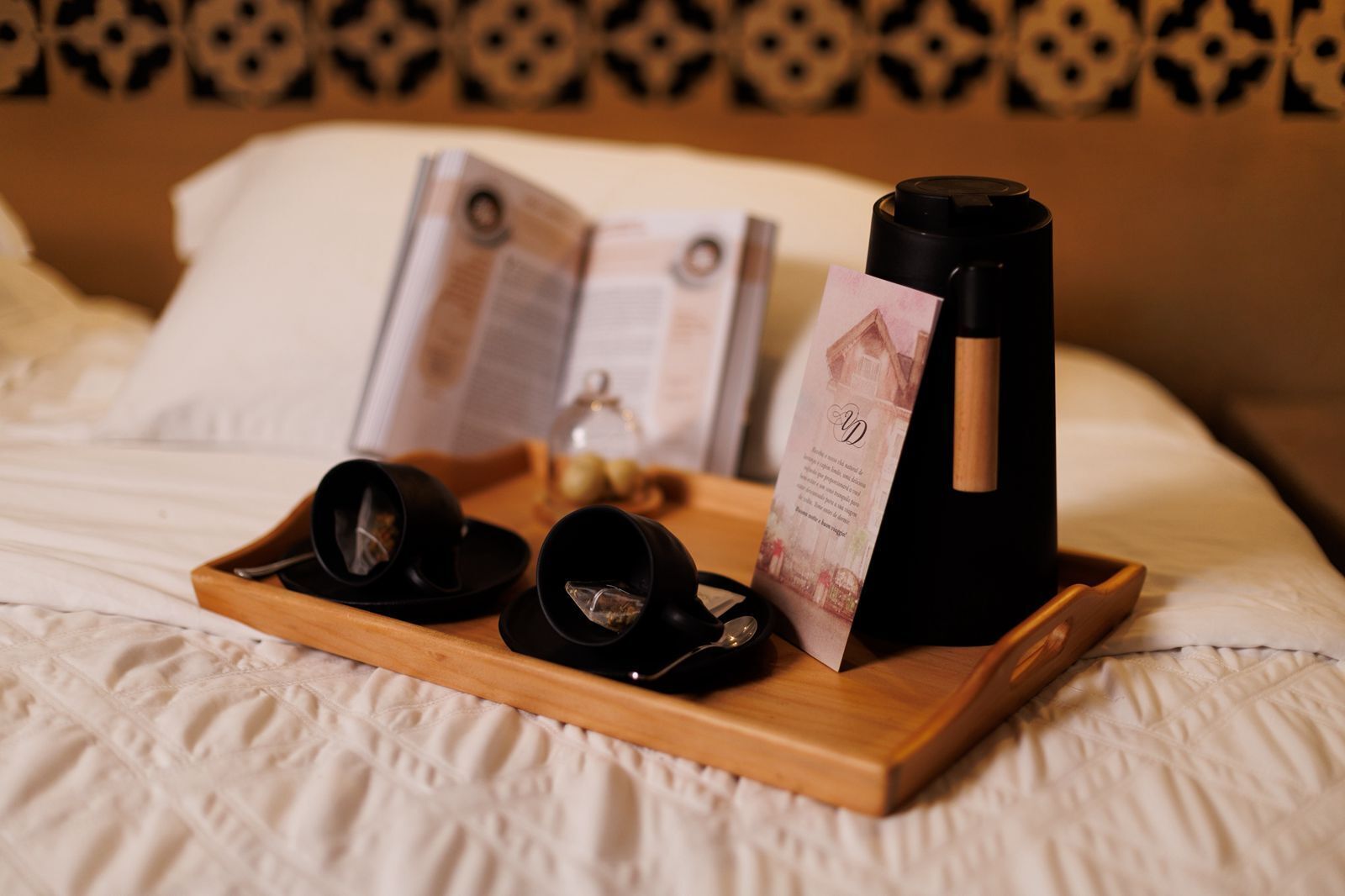 A bed with a wooden tray holding black cups, a wine bottle, and a menu.