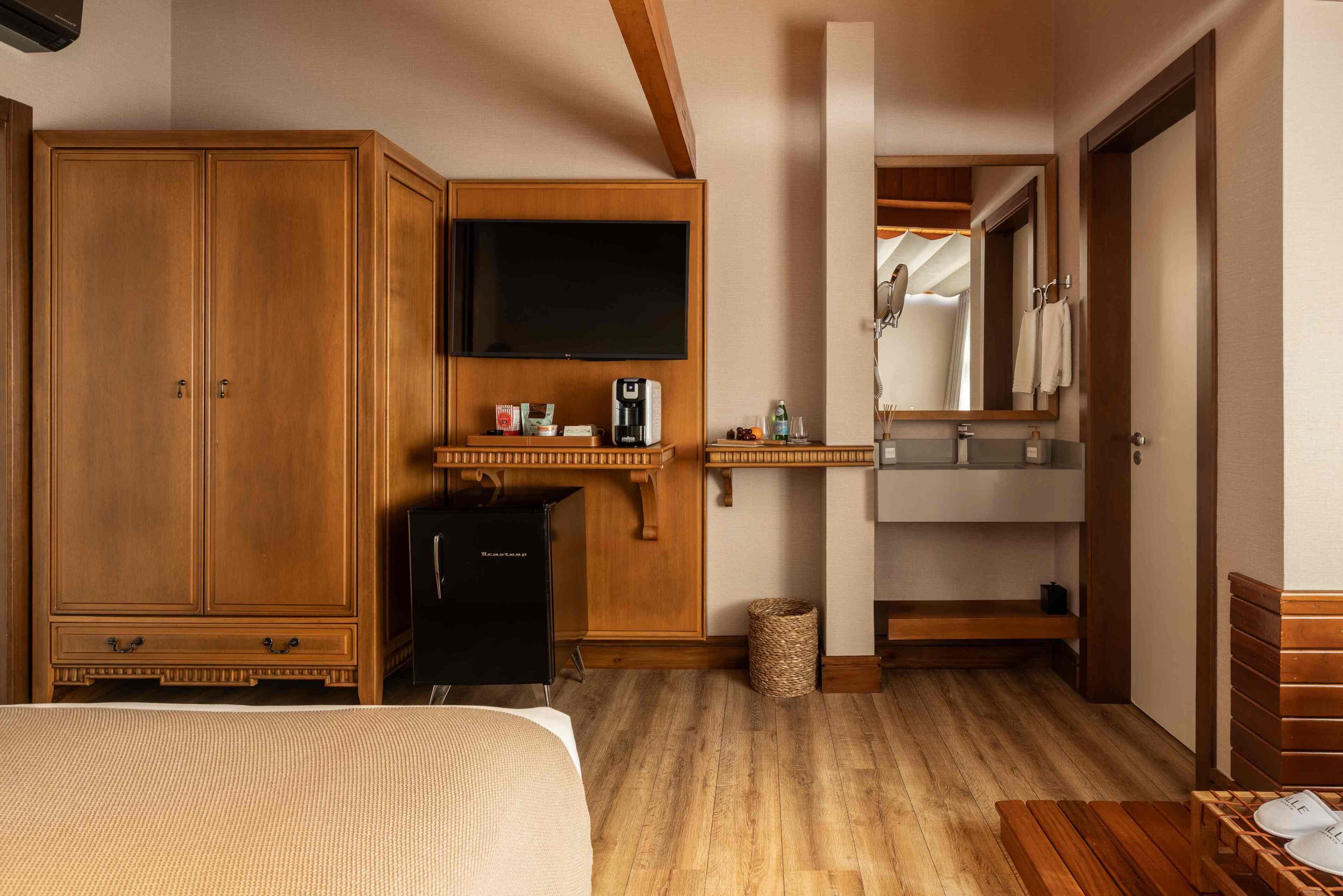 A hotel room with a large wardrobe, a flat-screen TV, a minibar, a wooden bench, and a mirror.