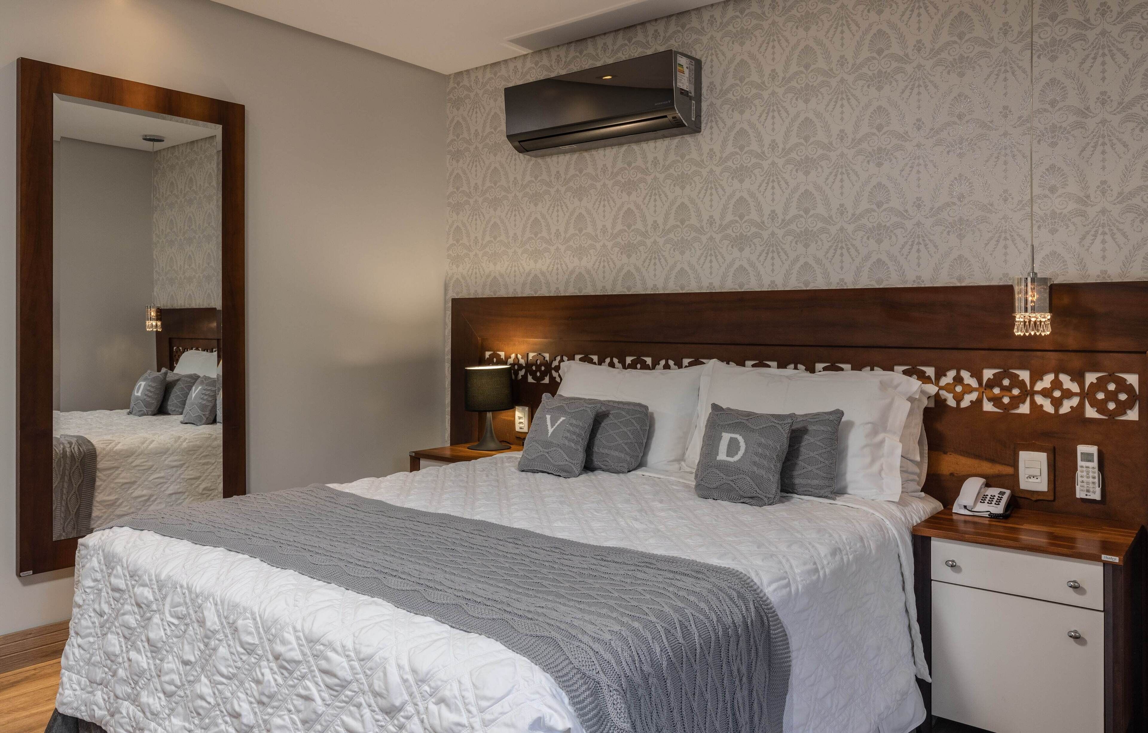 A hotel room with a large bed, a bedside table with a lamp, a mirror, a wall-mounted air conditioner, and a telephone.