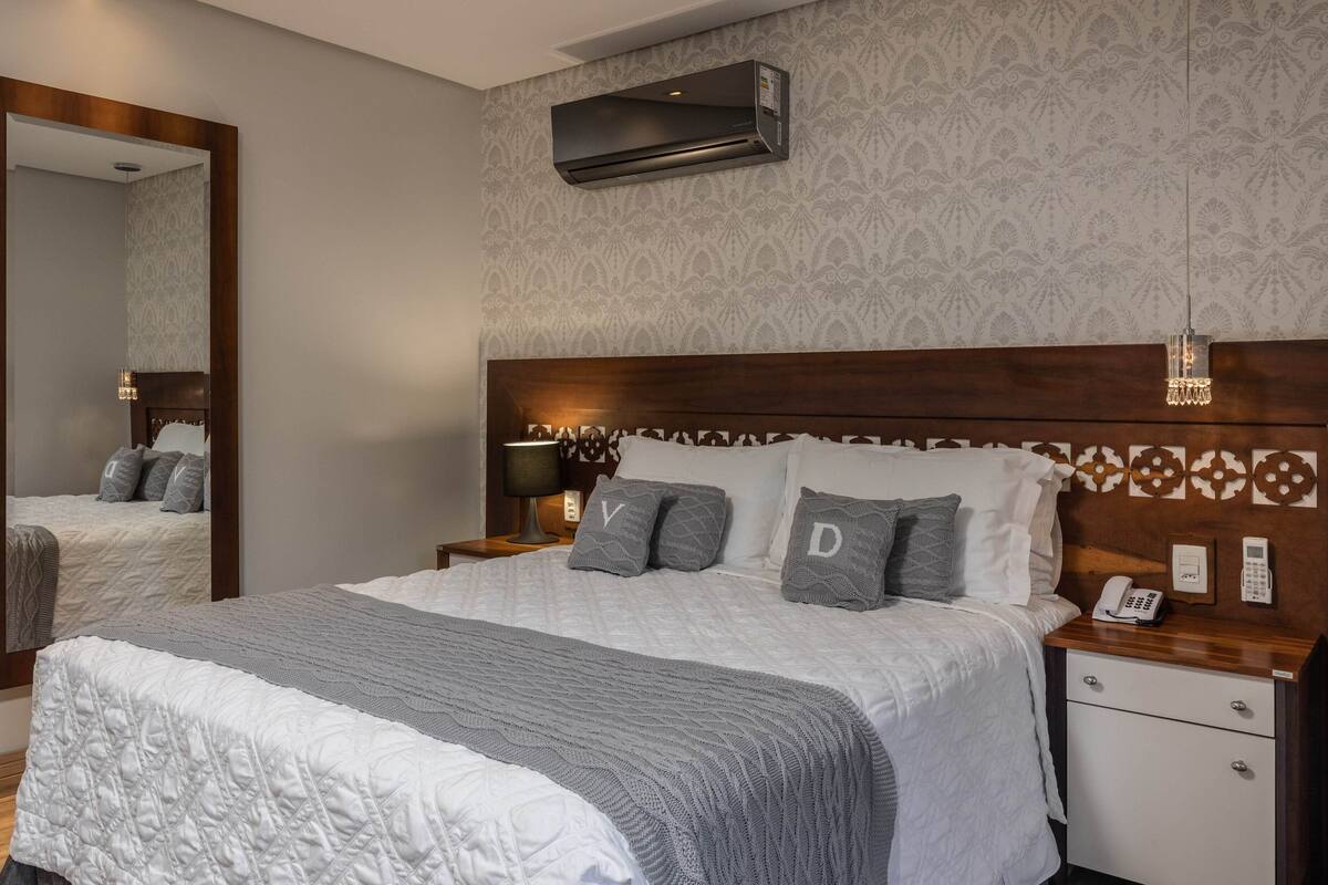 Habitación de hotel con una cama grande, una mesita de noche con lámpara, un espejo, un aire acondicionado instalado en la pared y un teléfono.