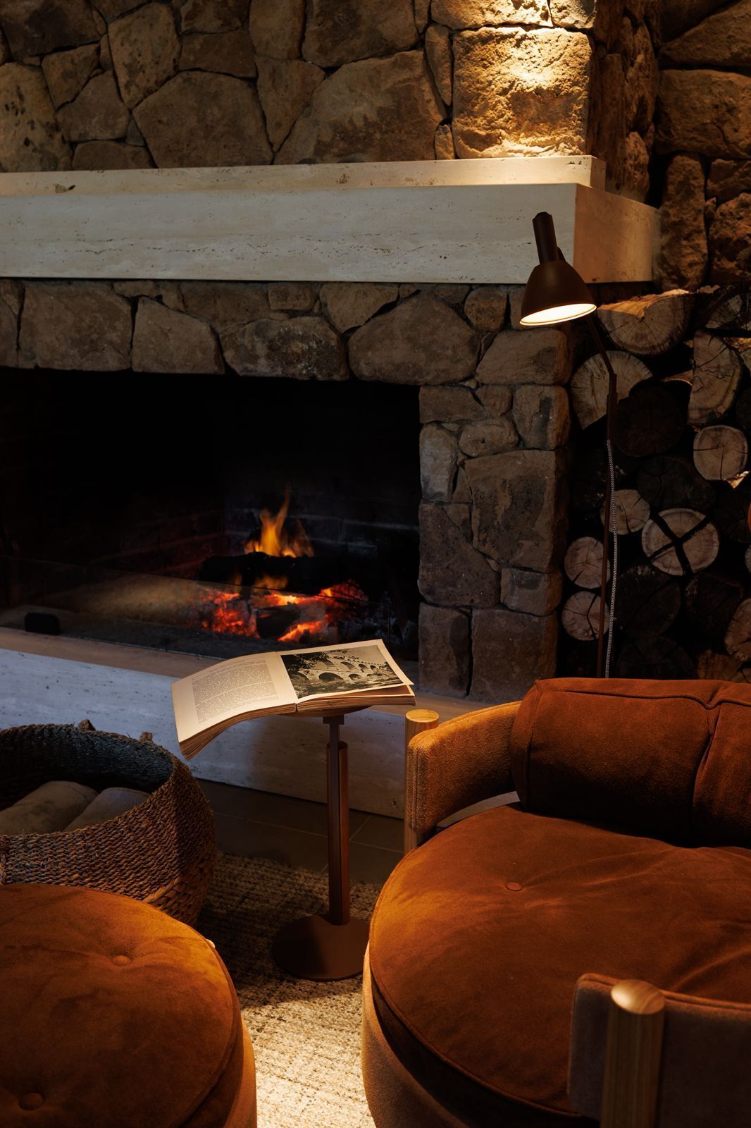 A stone fireplace with a lit fire, a book on a small table, and a brown sofa in a cozy room.