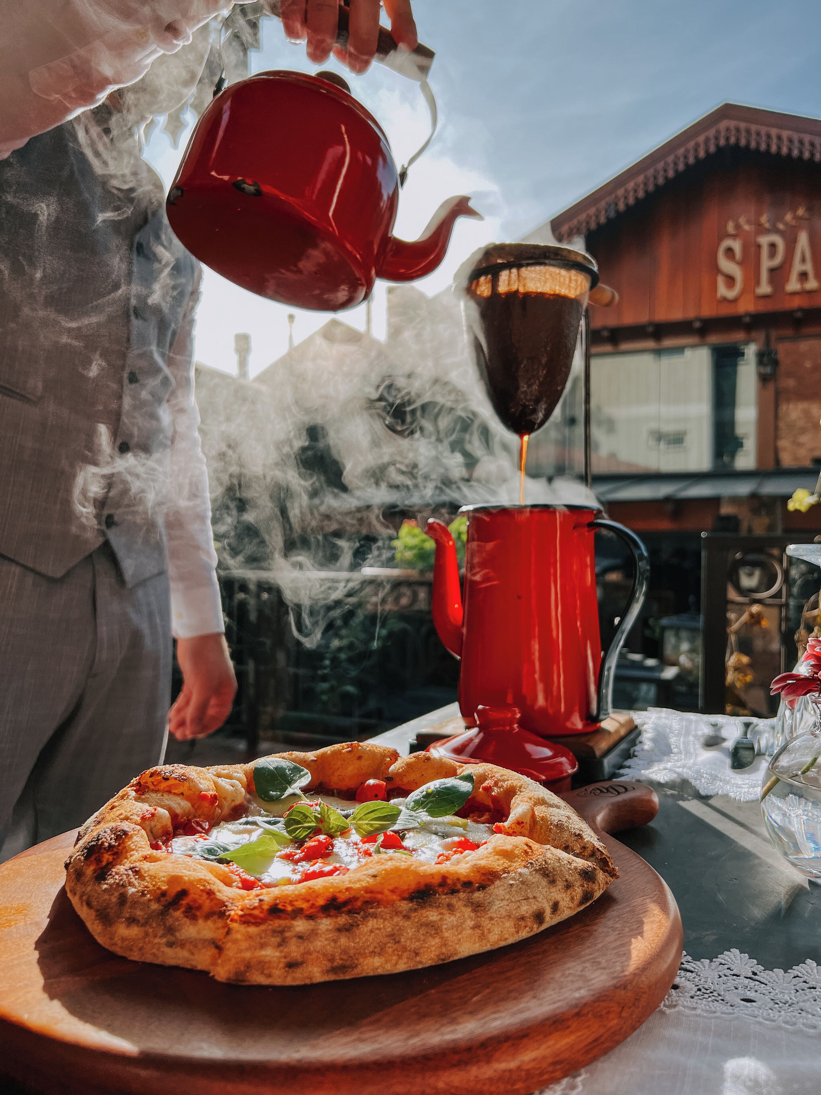 Una persona sirviendo una bebida caliente en una taza roja, con una pizza sobre una tabla de madera al frente.