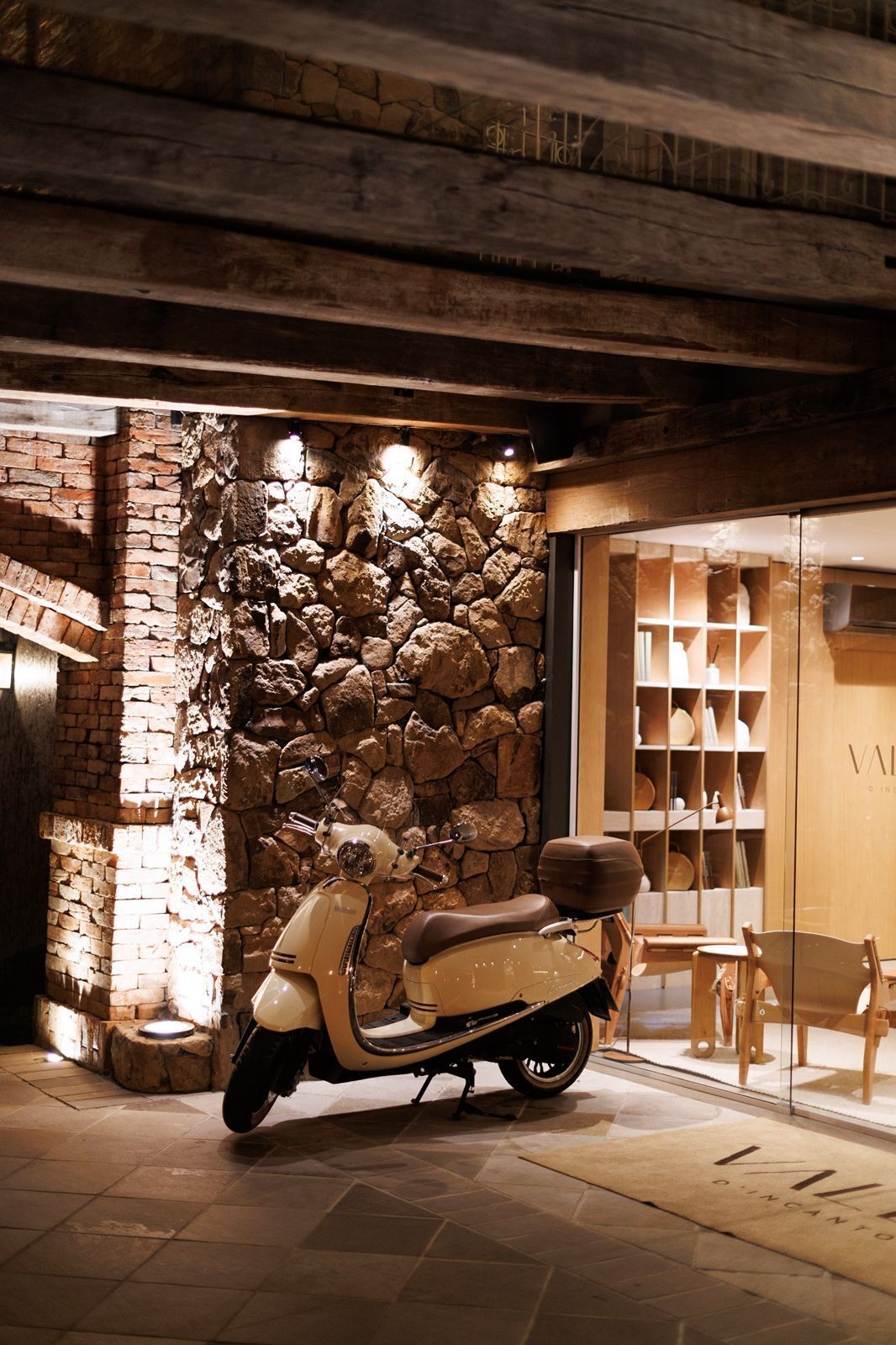 A vintage scooter parked in front of a stone wall with a wooden interior and a glass door leading to a room with shelves.
