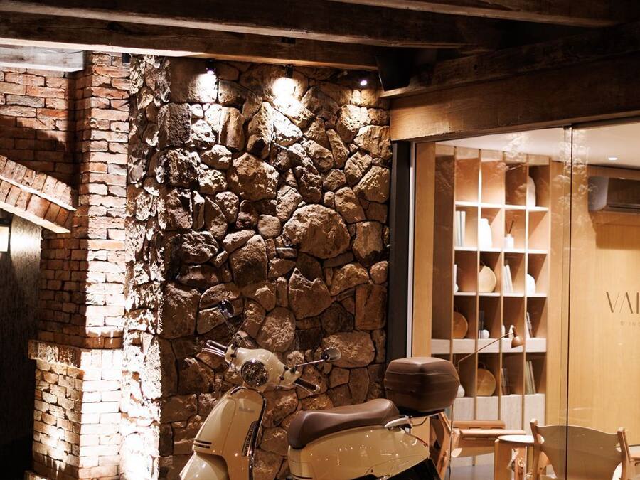 A vintage scooter parked in front of a stone wall with a wooden interior and a glass door leading to a room with shelves.
