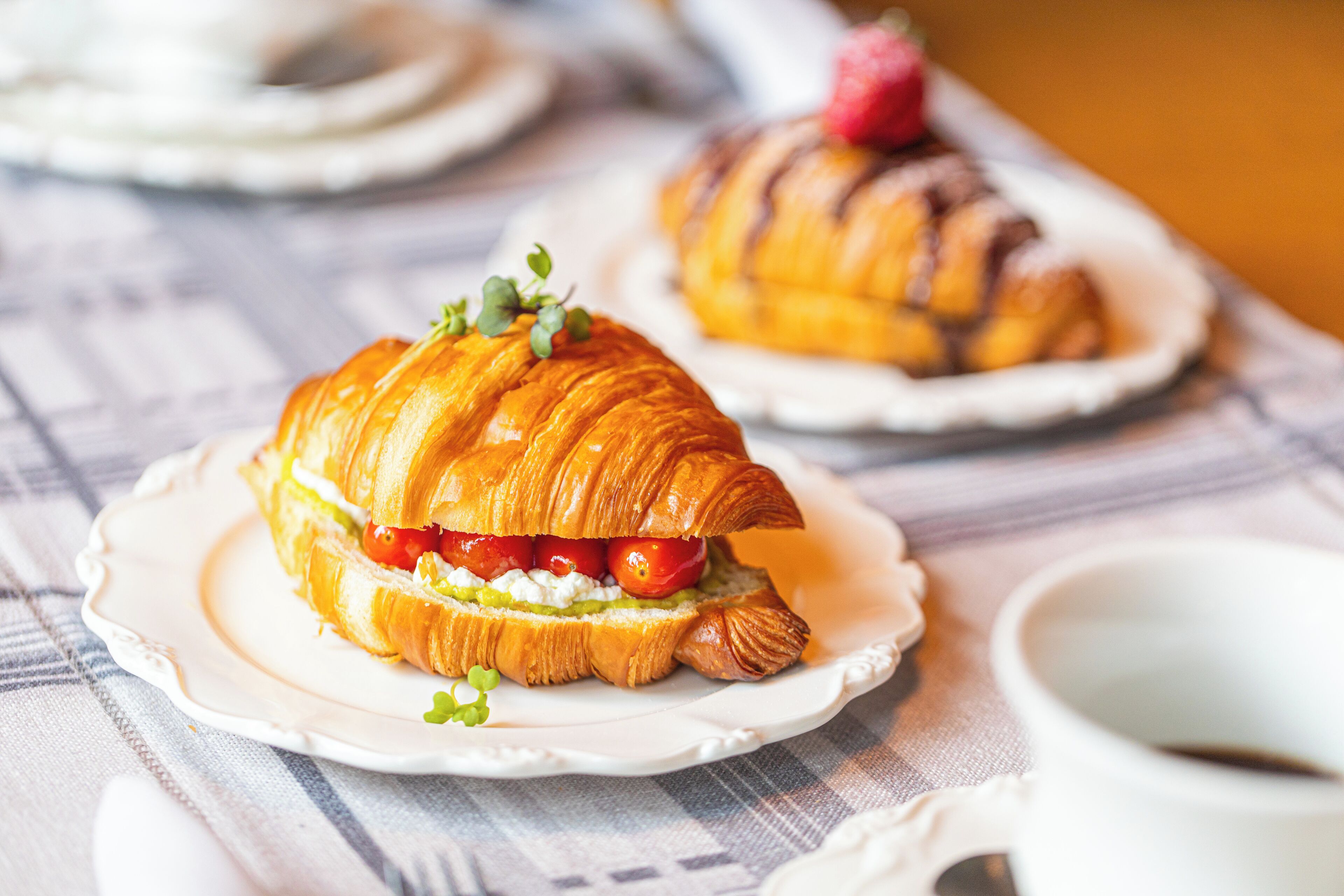 Sándwich de croissant sobre un plato blanco, con una taza al fondo.