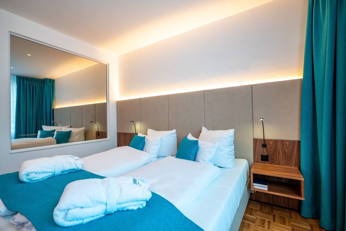 Habitación de hotel con dos camas, un espejo grande, una mesita de noche con lámpara y un cabecero de madera.
