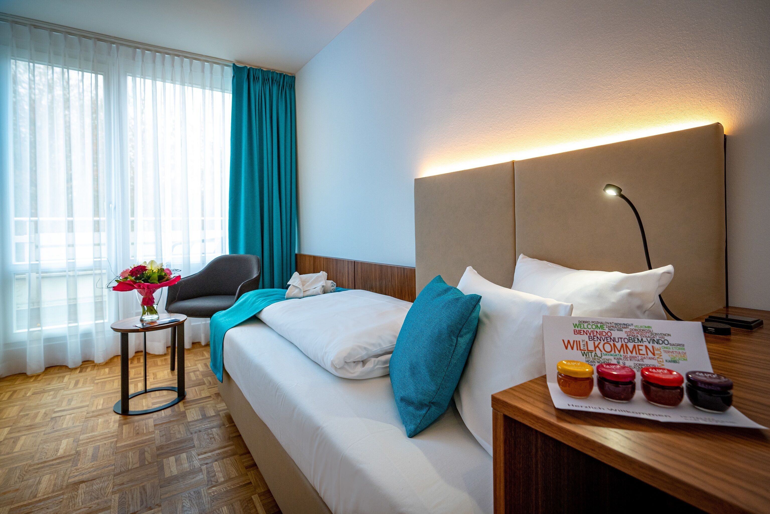 A hotel room with a bed, a nightstand with a lamp, a chair, a small table with a vase of flowers, and a window with curtains.