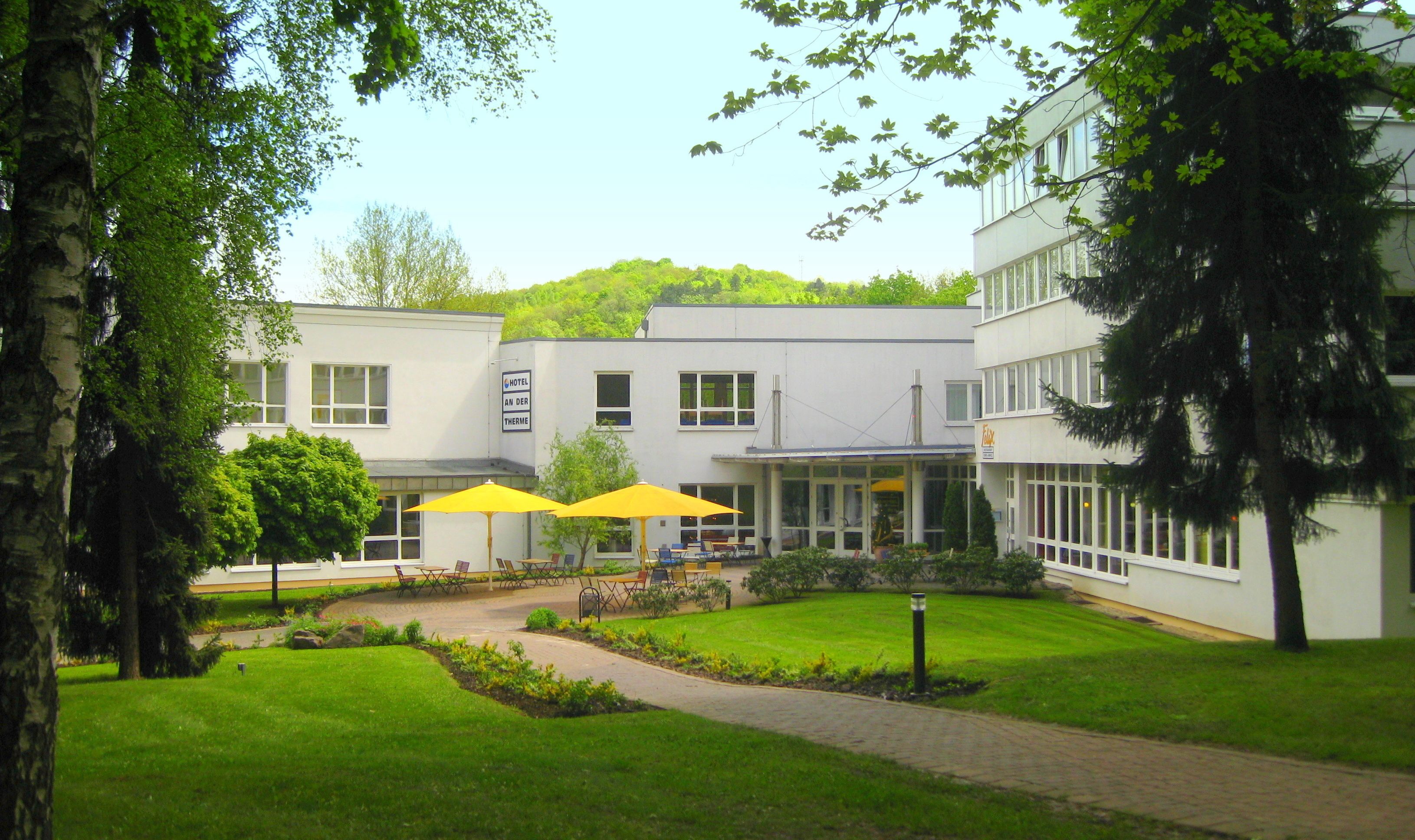 A hotel courtyard with outdoor seating, a pathway, and a building with a sign reading "HOTEL AN DER THERME".