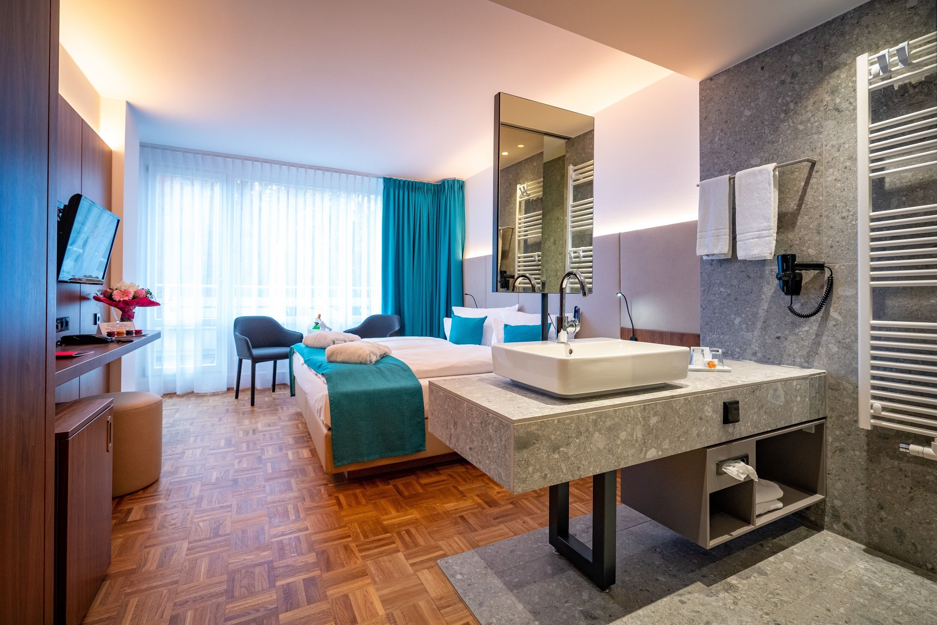 A modern hotel room with a bed, a desk with a computer, a large mirror, a sink, and a shower area.