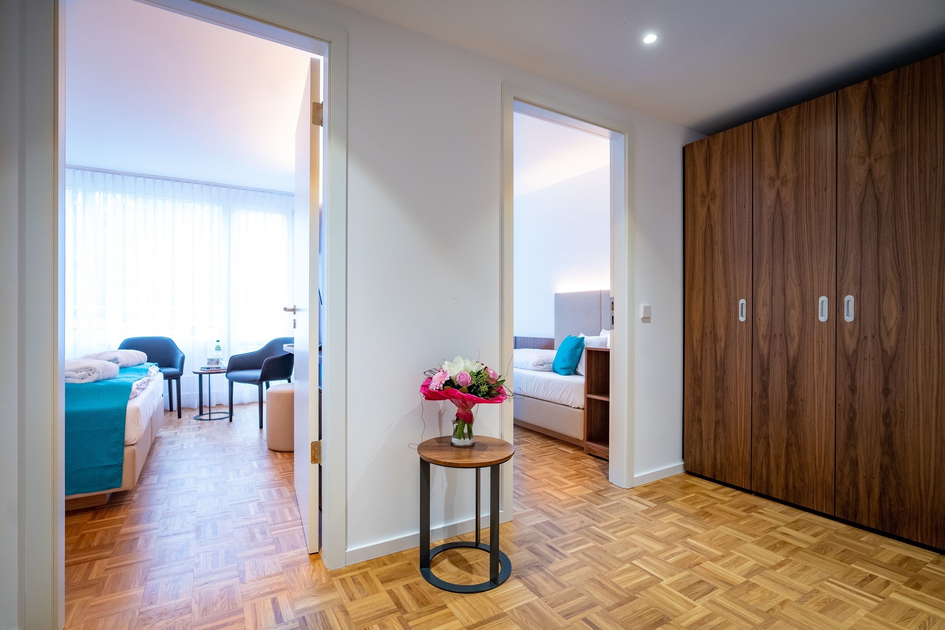 A hotel room with a wooden floor, a bed with a blue cover, a small round table with a vase of flowers, and a wardrobe.