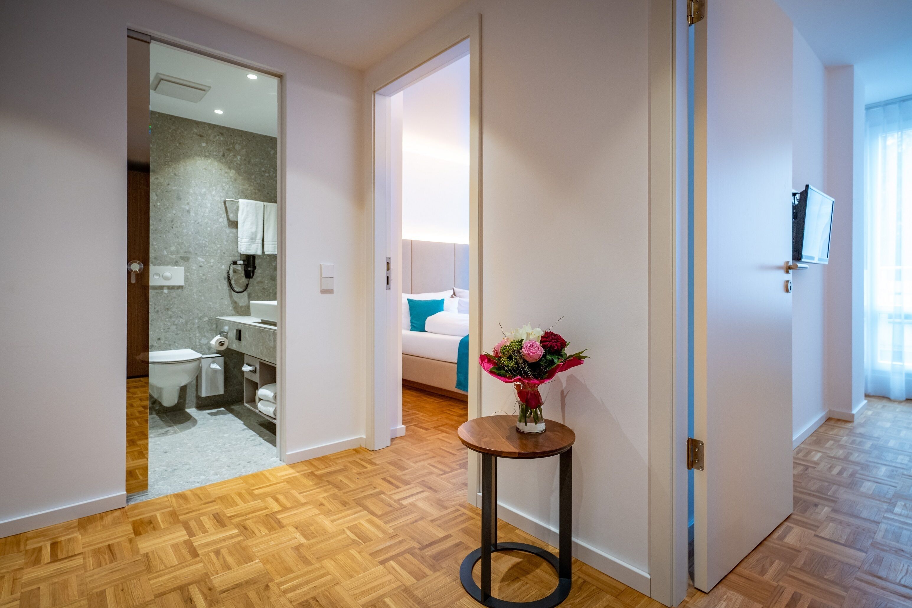 Ein Hotelzimmer mit Holzboden, einem Badezimmer mit Dusche und Toilette, einem Bett mit blauem Kopfkissen und einer Vase mit Blumen auf einem kleinen Tisch.