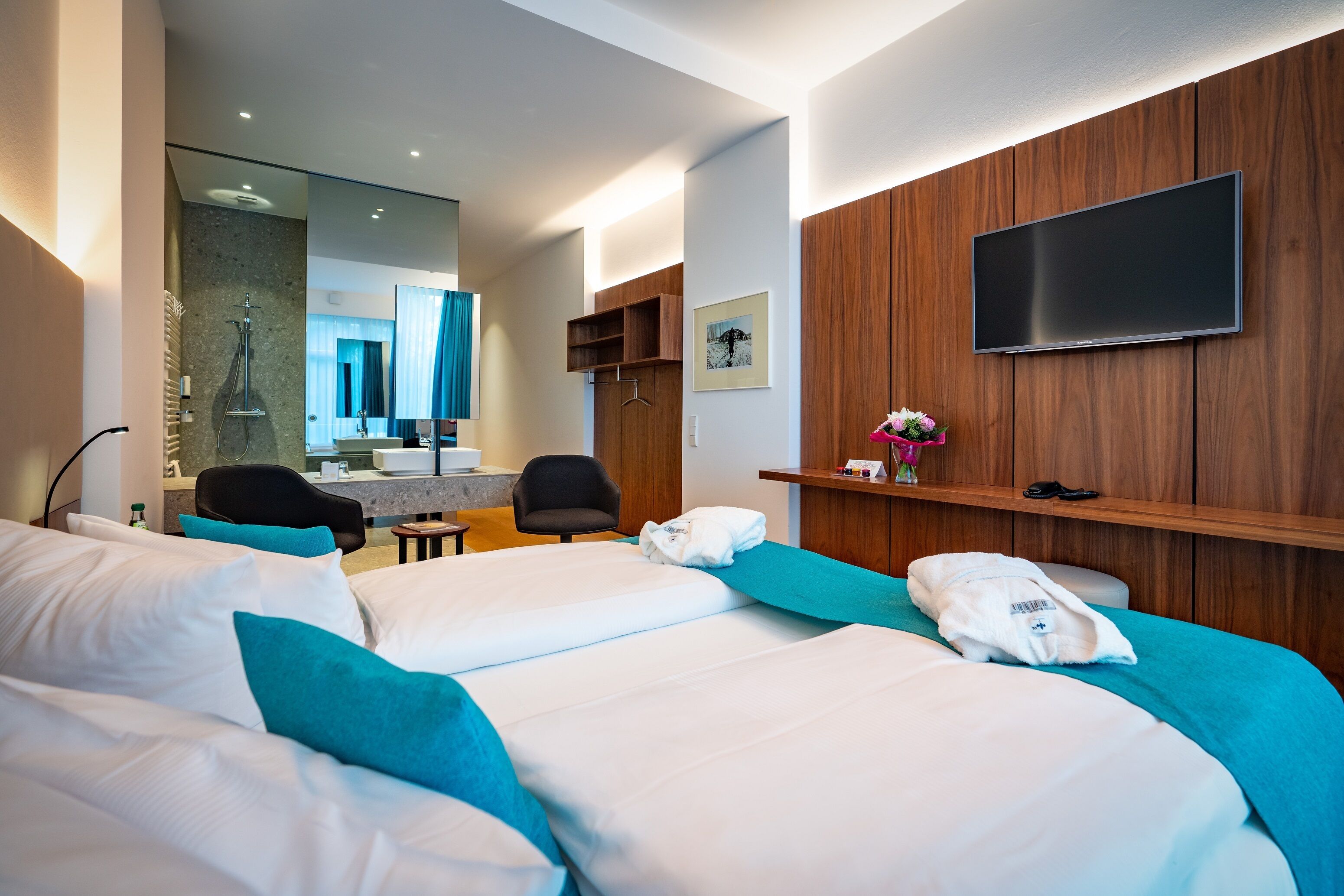 Una habitación de hotel moderna con una cama grande, un televisor montado en la pared, un diseño de paneles de madera y un baño visible a través de una puerta abierta.