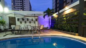 Zona de piscina con sillones de descanso y sombrillas, rodeada de edificios y un muro decorativo con plantas.