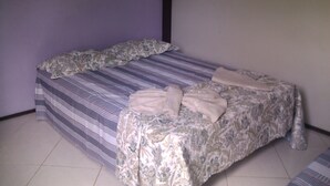 A single bed with a floral-patterned bedspread and a striped blanket, positioned against a plain wall with a tiled floor. - Flat do Indio (Fernando de Noronha)