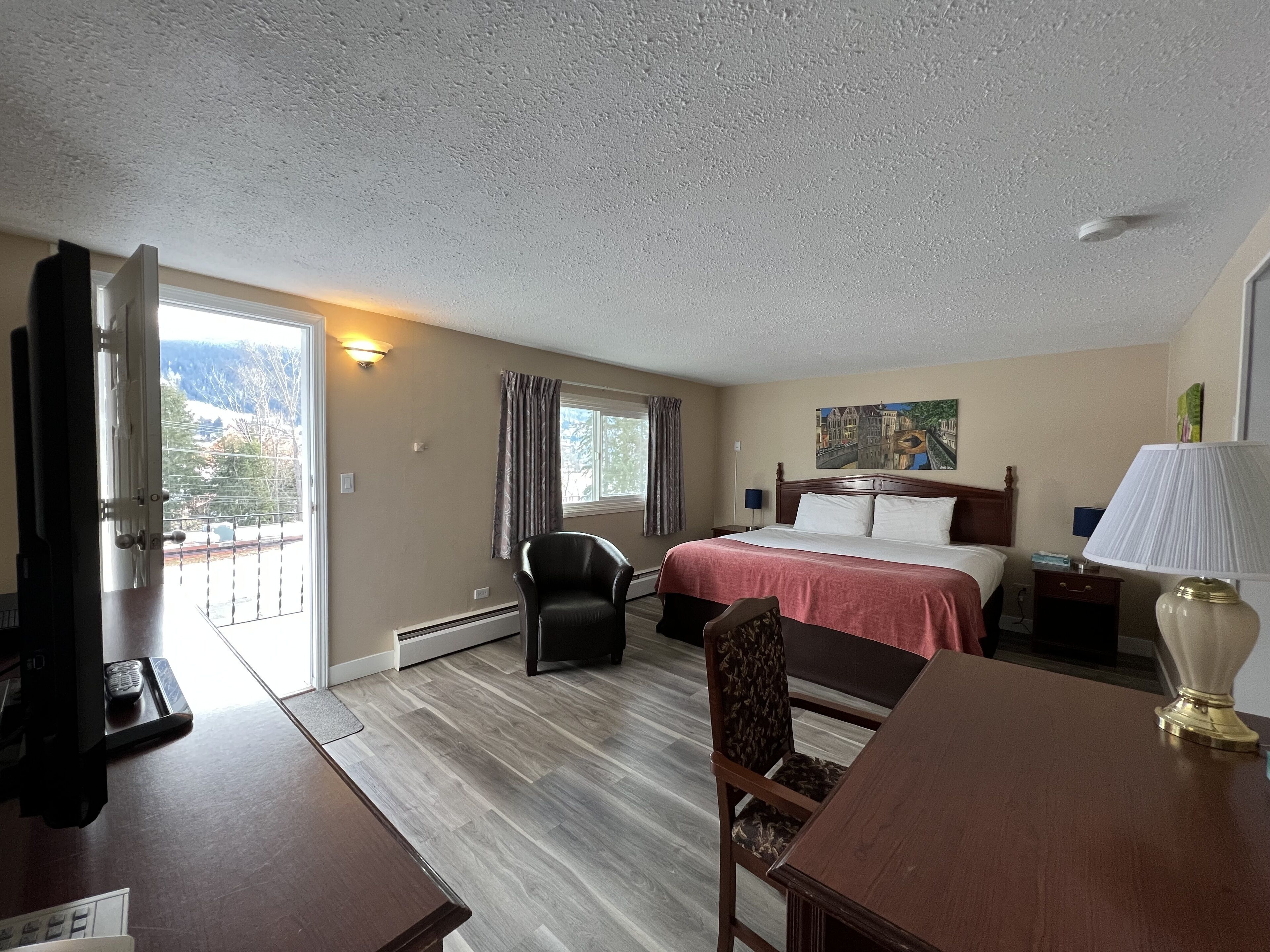 A hotel room with a bed, a desk, a chair, a lamp, a painting, and a view of the outdoors.