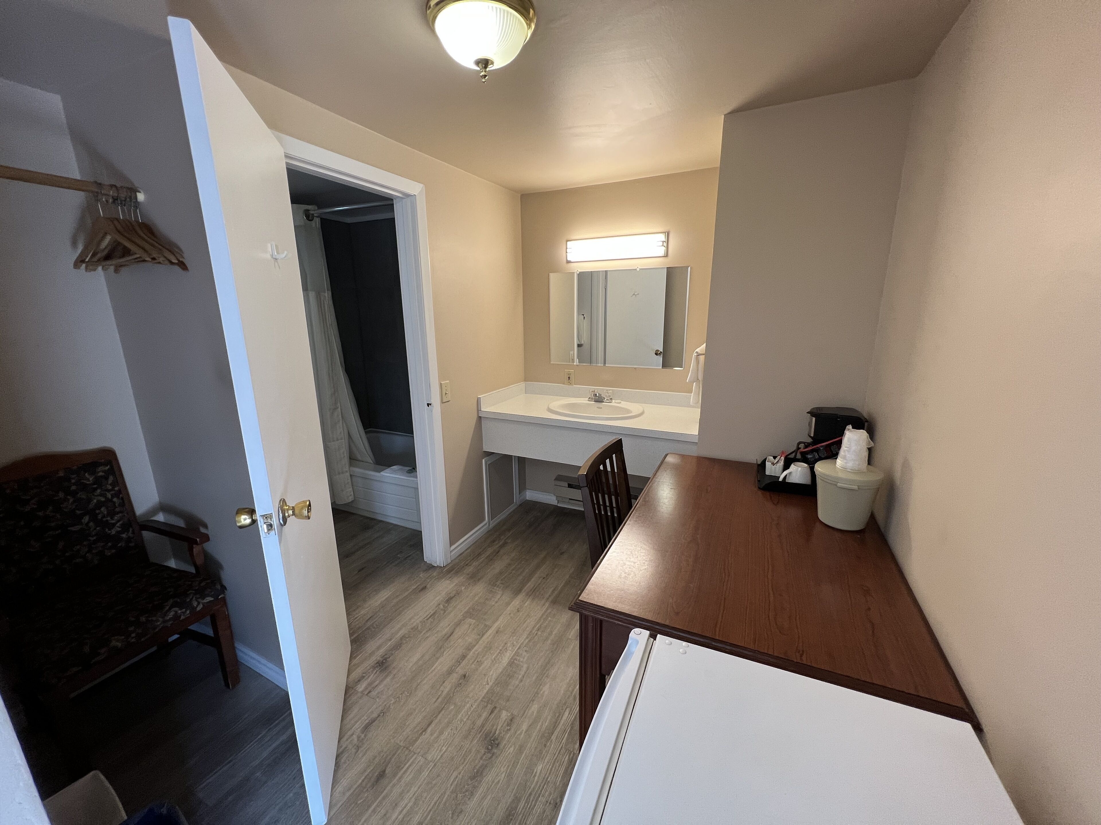 Une salle de bain d’hôtel avec un lavabo, un miroir et des toilettes. On y trouve une chaise et une table sur laquelle se trouve une machine à café.