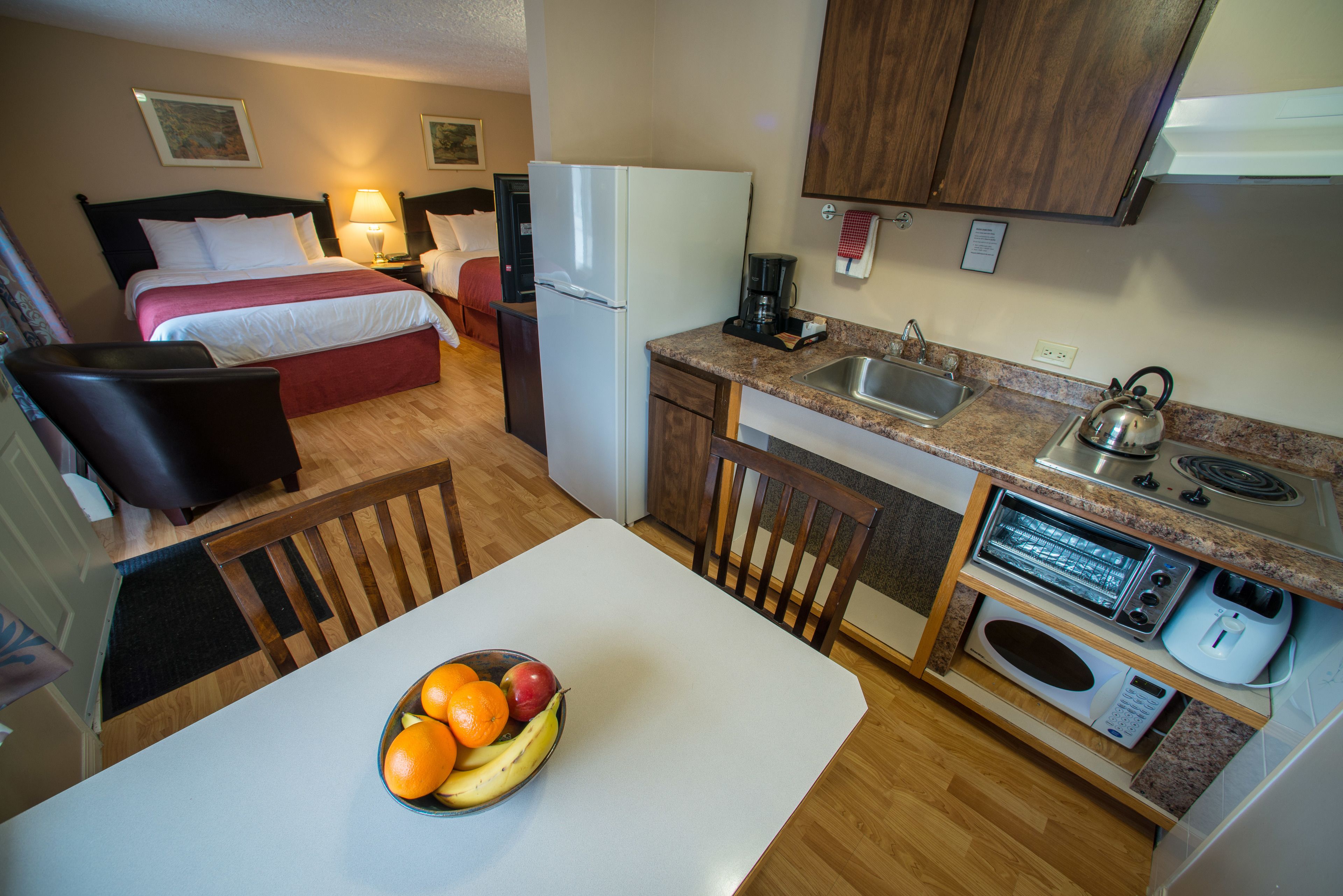 Une chambre d’hôtel compacte comprenant un lit, une petite kitchenette, un coin repas avec une table et des chaises, et un panier de fruits sur la table.