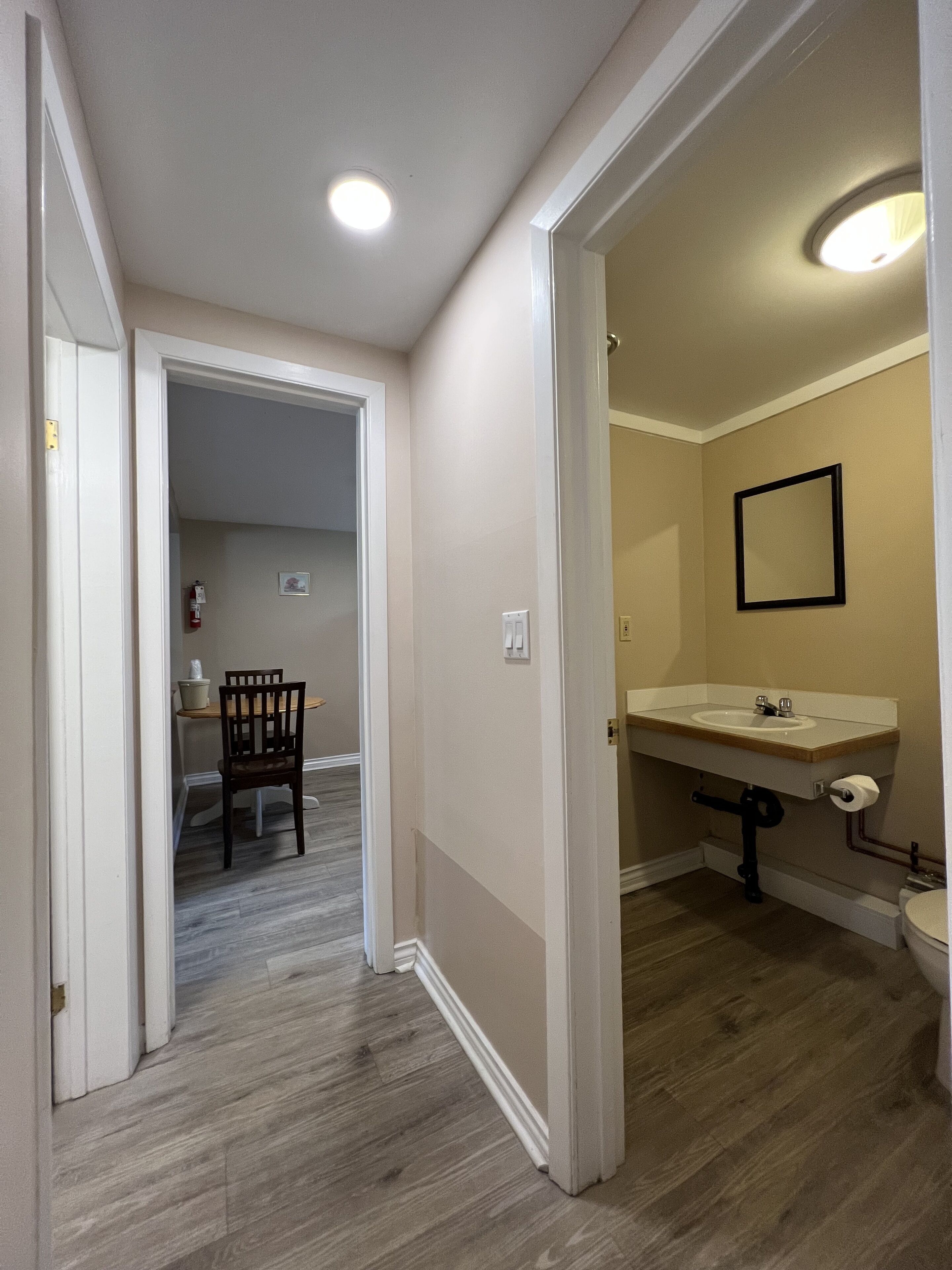 A hallway leading to a bathroom with a sink and mirror, a chair in the adjacent room, and a door on the left.