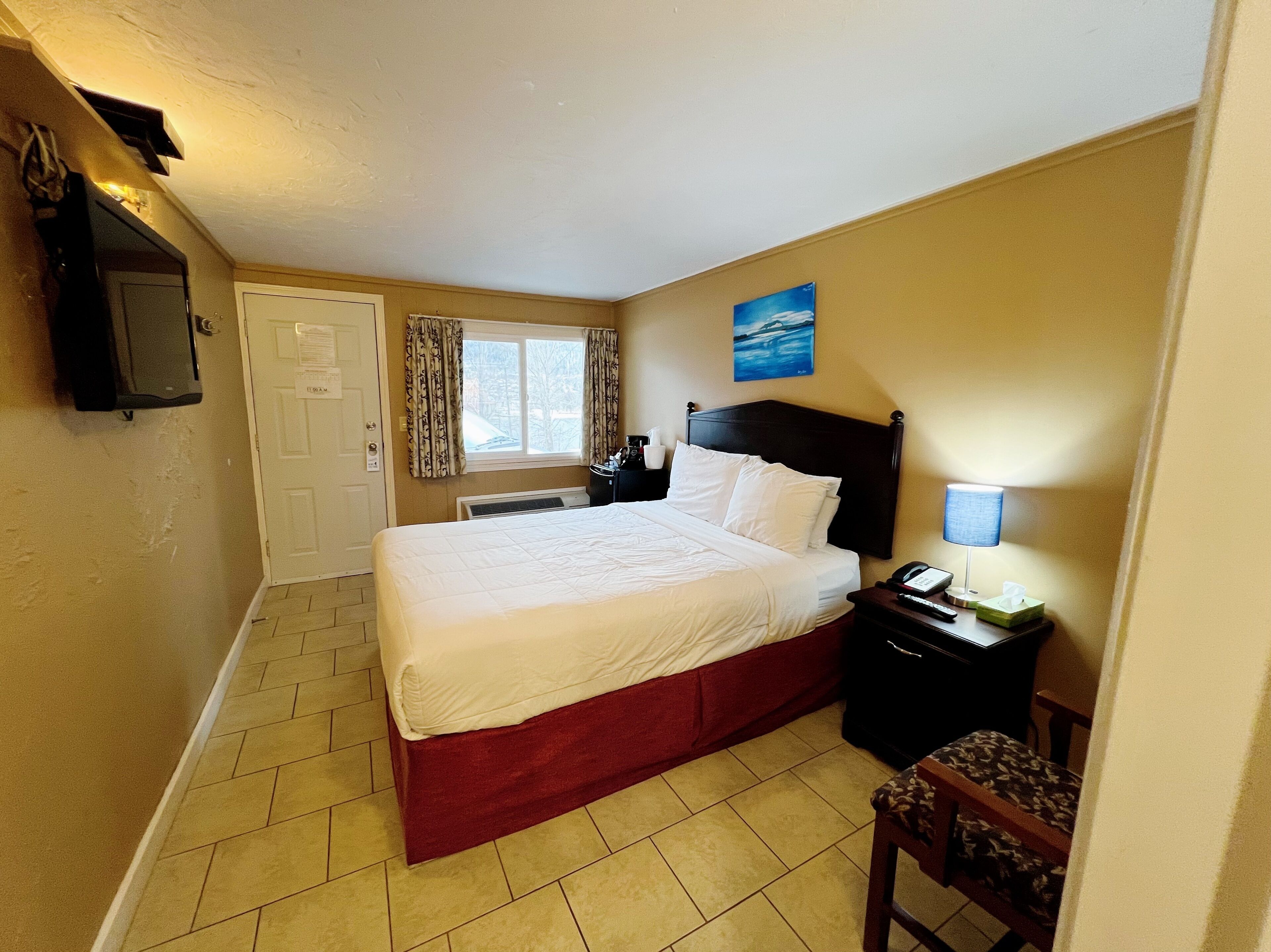 A hotel room with a bed, a nightstand, a chair, a window with curtains, a wall-mounted TV, and a painting.