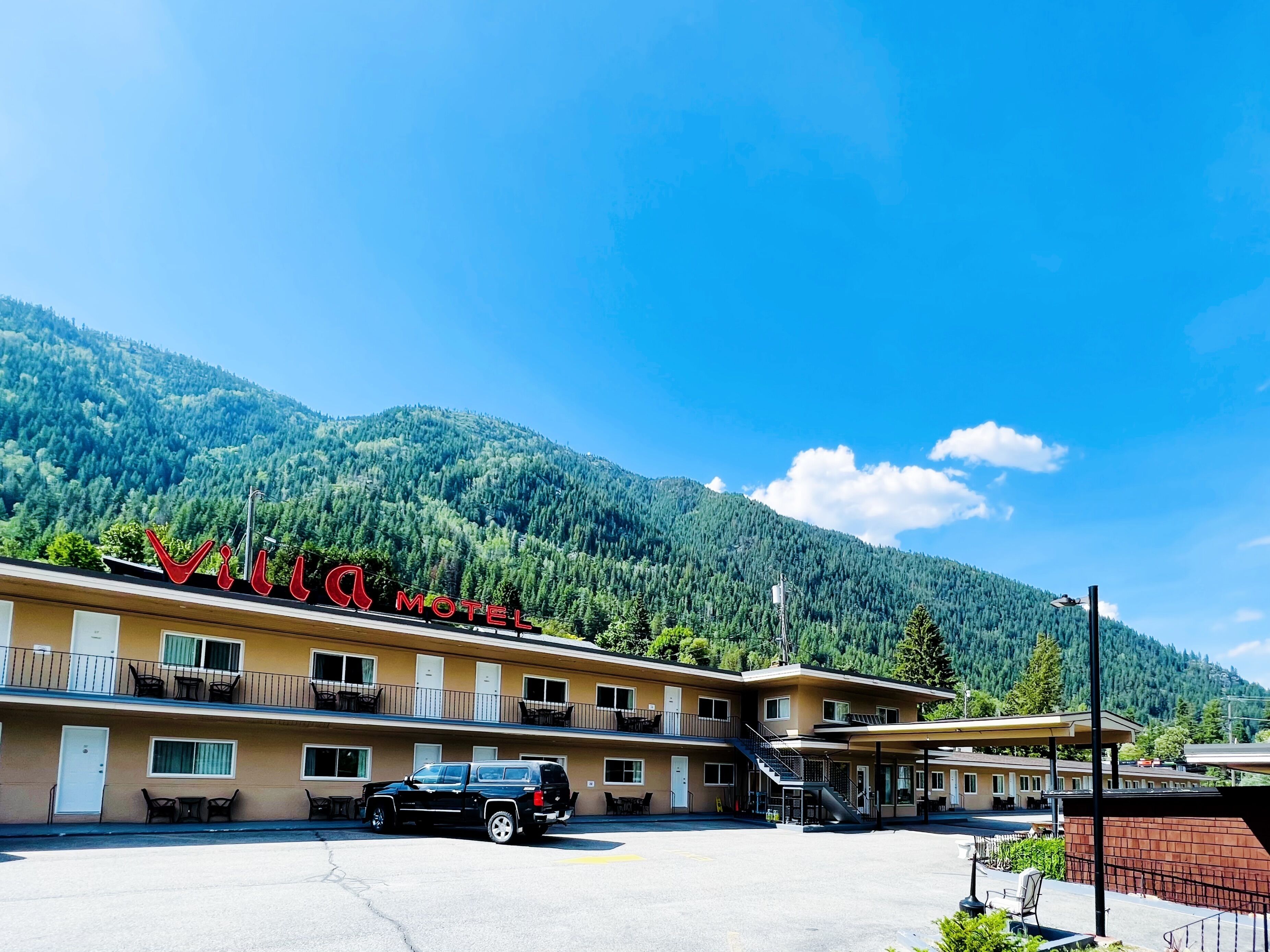 A motel with a mountain backdrop, featuring a parking lot and a black SUV parked in front.