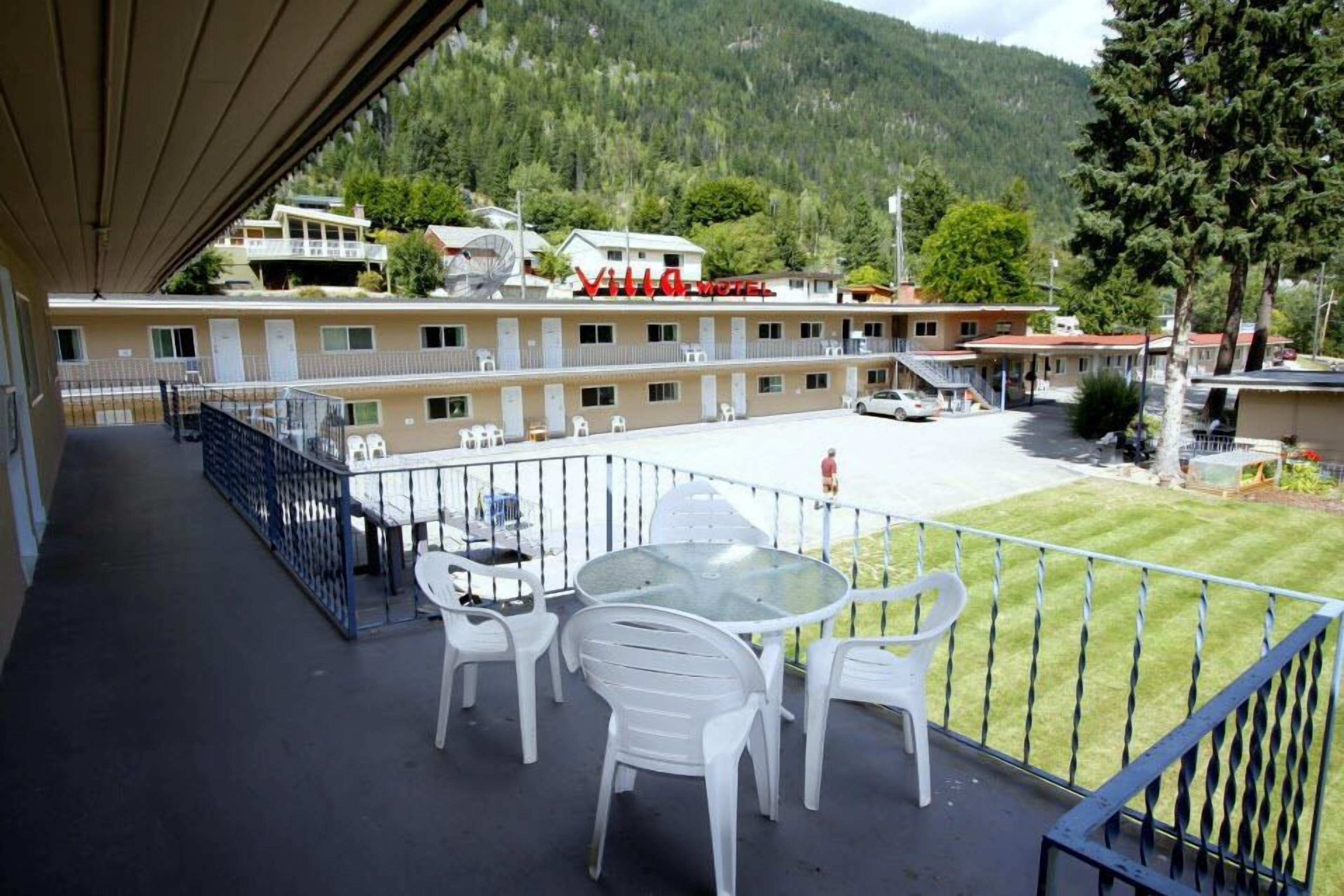 A hotel with a balcony featuring a glass table and white plastic chairs, overlooking a parking area and a mountainous landscape.