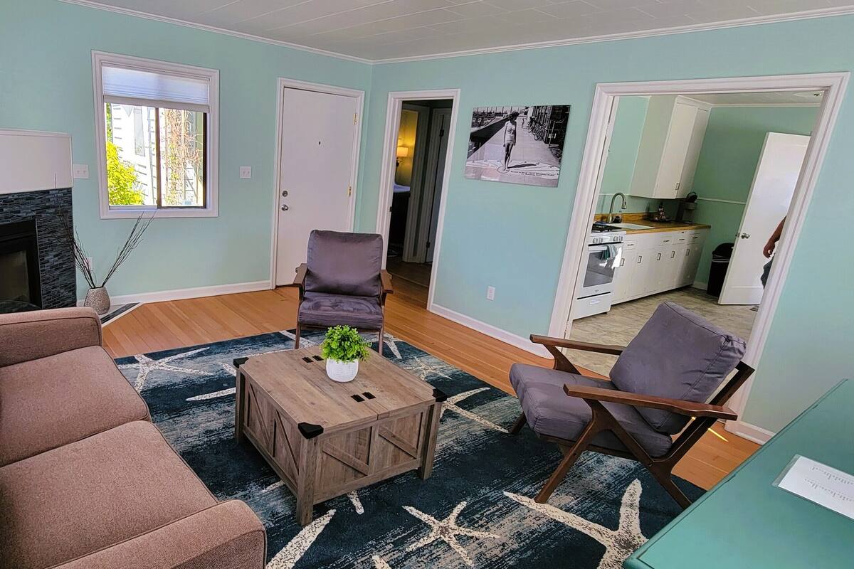 A living room with a sofa, coffee table, and chairs. There is a fireplace, a window, and a kitchen visible through an open door.