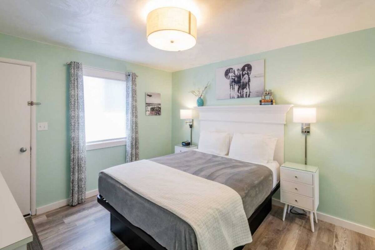 A bedroom with a bed, bedside tables, a nightstand, a window with curtains, a wall-mounted artwork, and a ceiling light fixture.