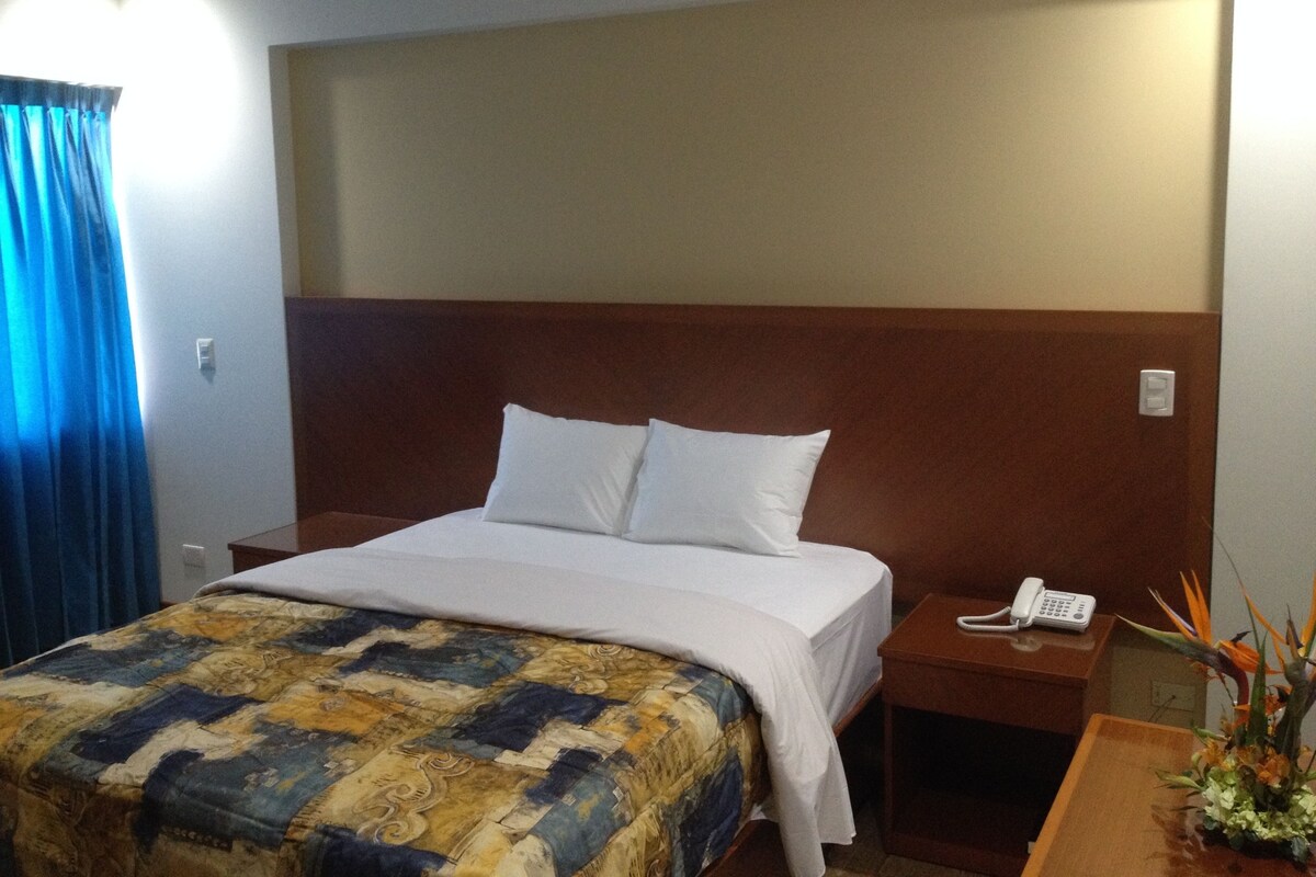 A hotel room with a bed, a nightstand with a phone, a wooden headboard, and a window with curtains.