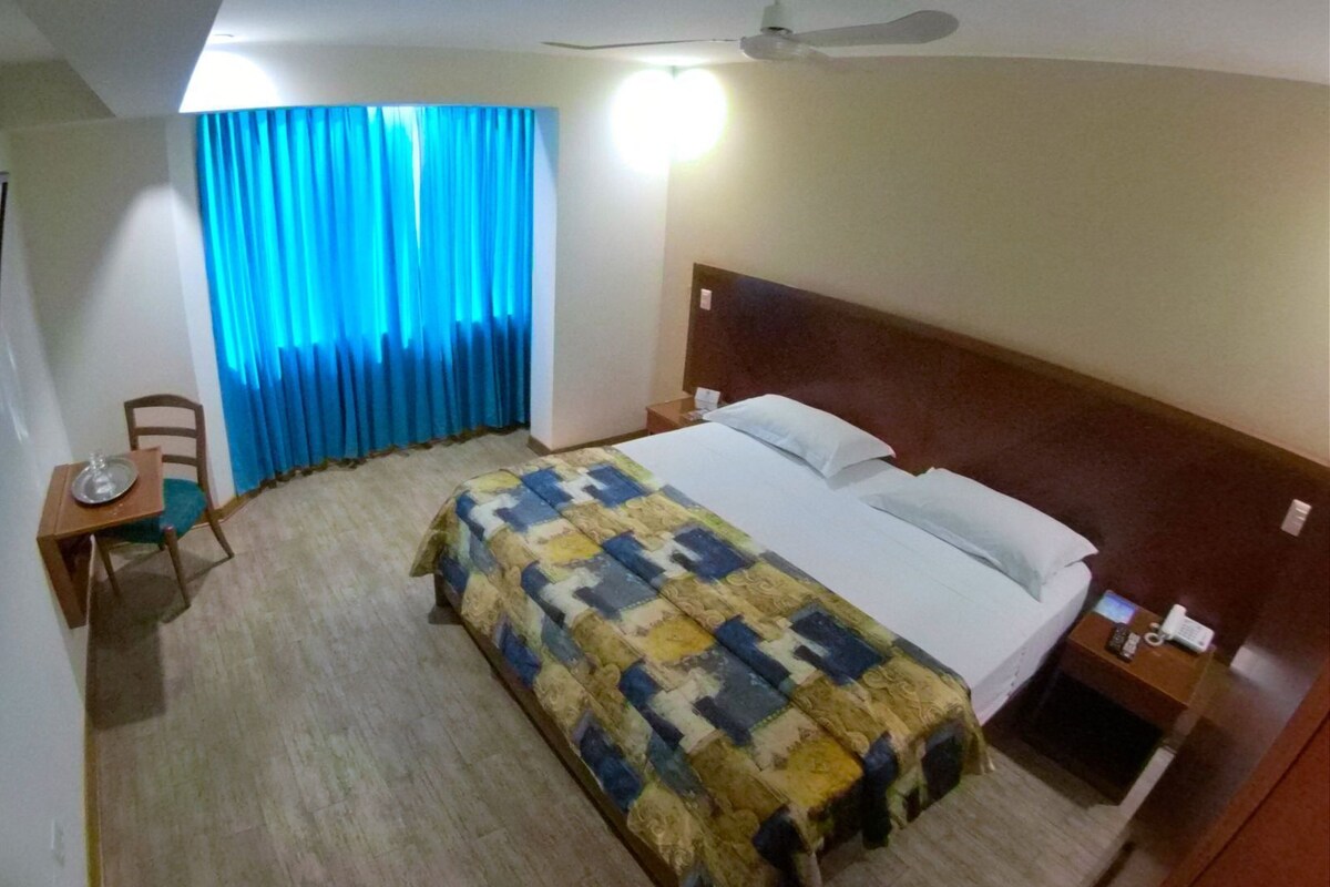 A hotel room with a bed, a desk with a chair, a mirror, and a window with curtains.