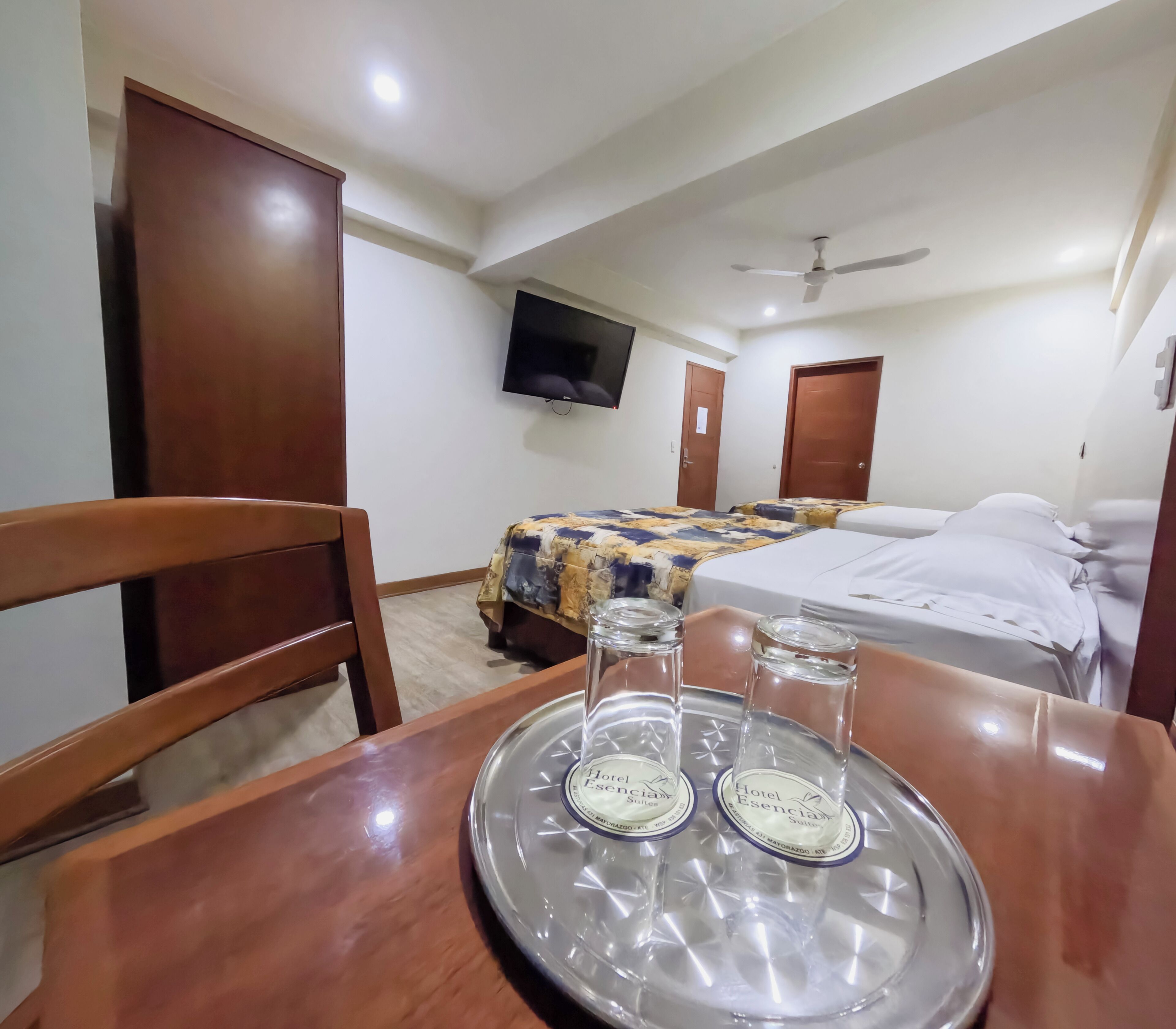 Habitación de hotel con cama, televisor montado en la pared y mesita con bandeja que contiene dos vasos.