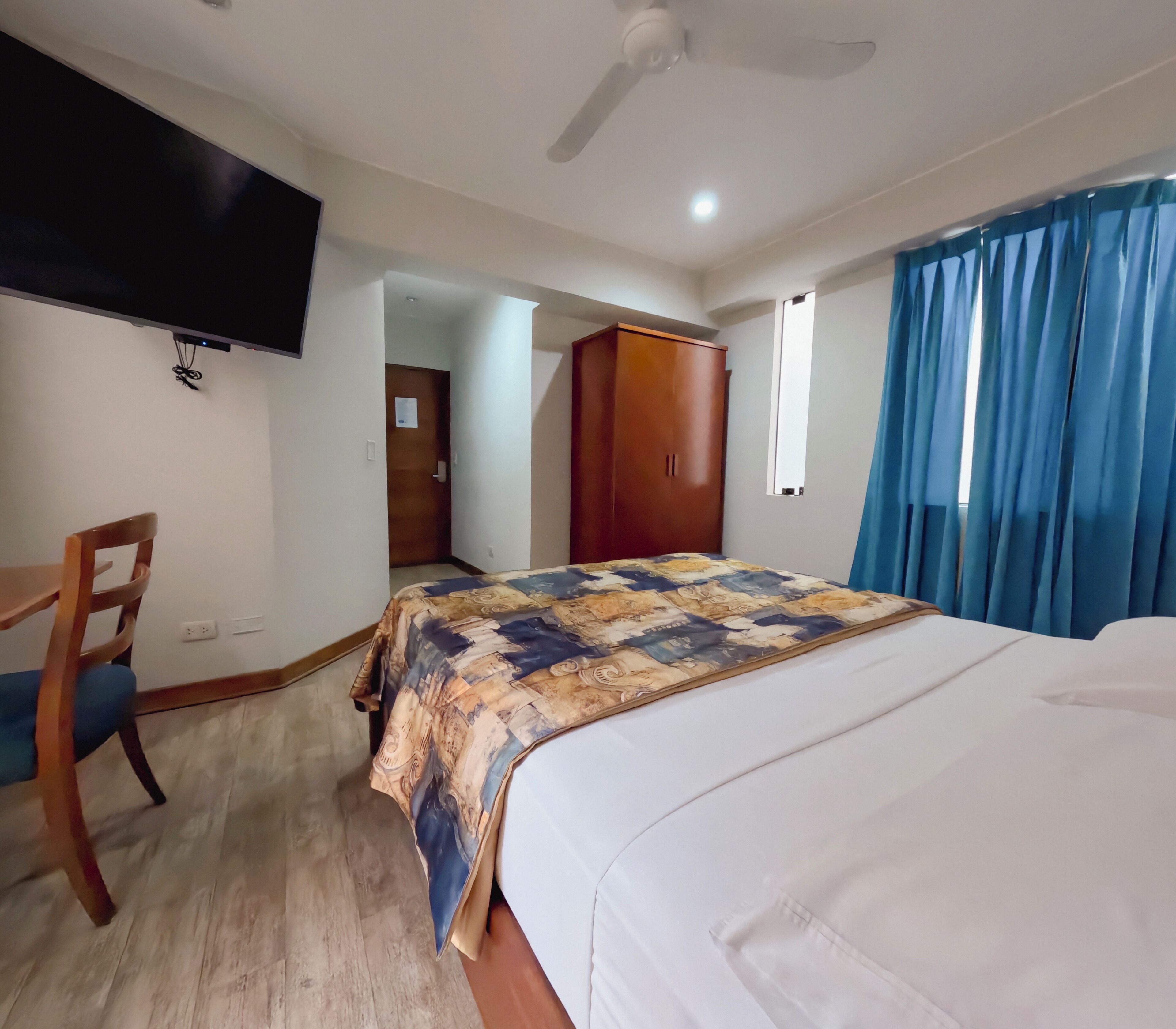 A hotel room with a bed, a desk, a chair, a wardrobe, a television mounted on the wall, and a ceiling fan.