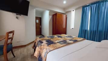 A hotel room with a bed, a desk, a chair, a wardrobe, a television mounted on the wall, and a ceiling fan.