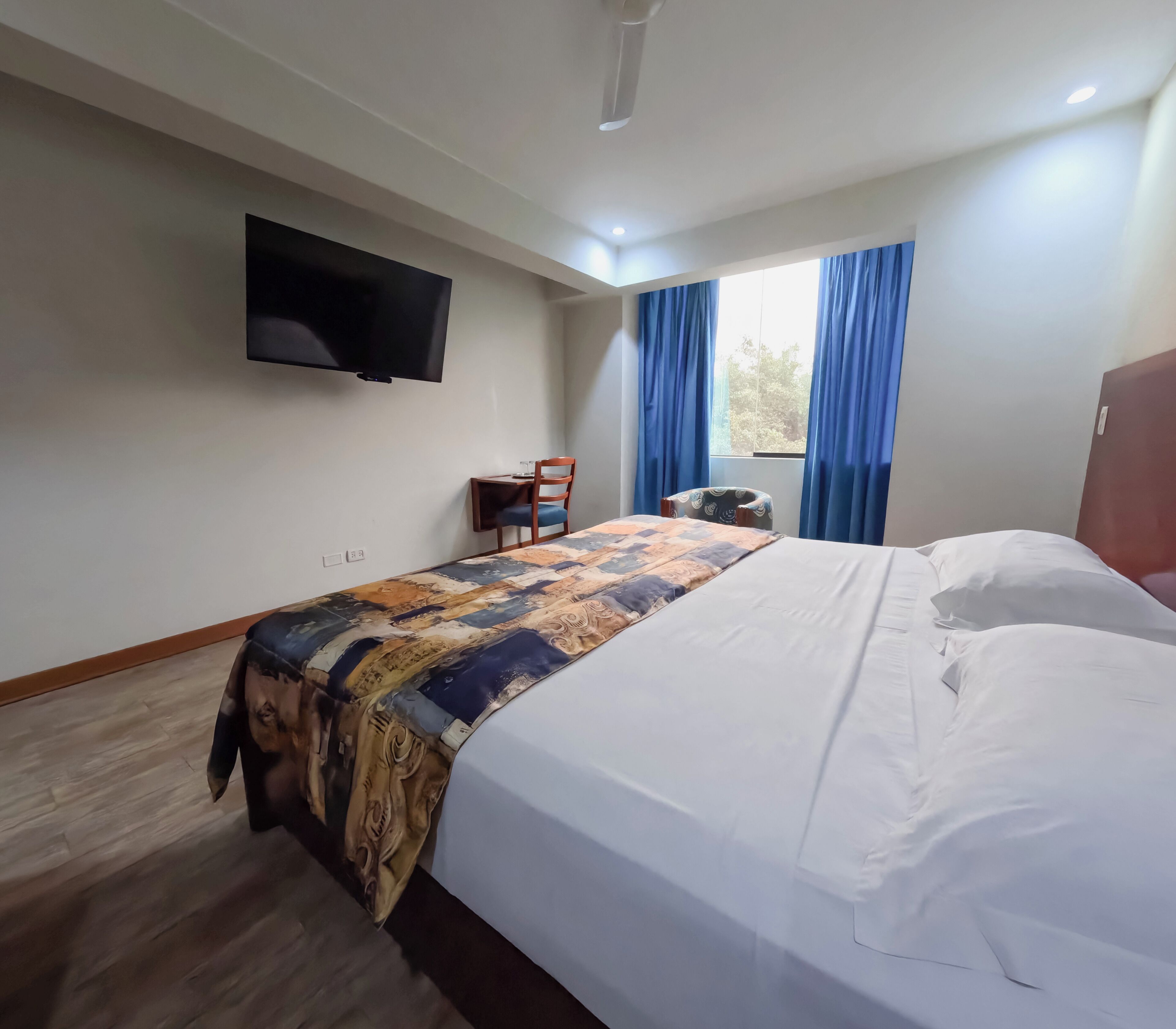 Quarto de hotel com cama, televisão de tela plana na parede, uma mesinha com cadeiras e uma janela com cortinas.