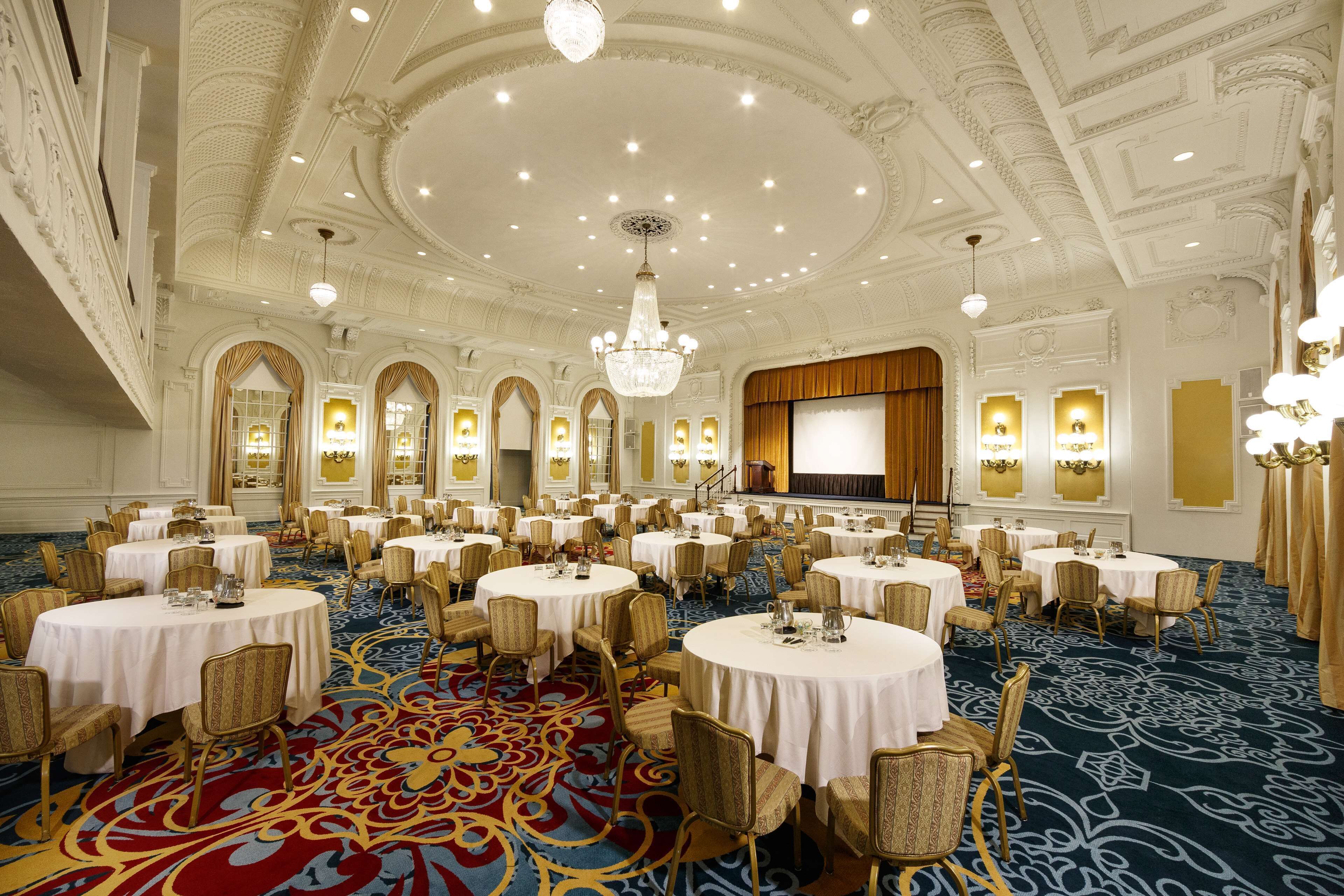 A large banquet hall with round tables, chairs, and a stage.
