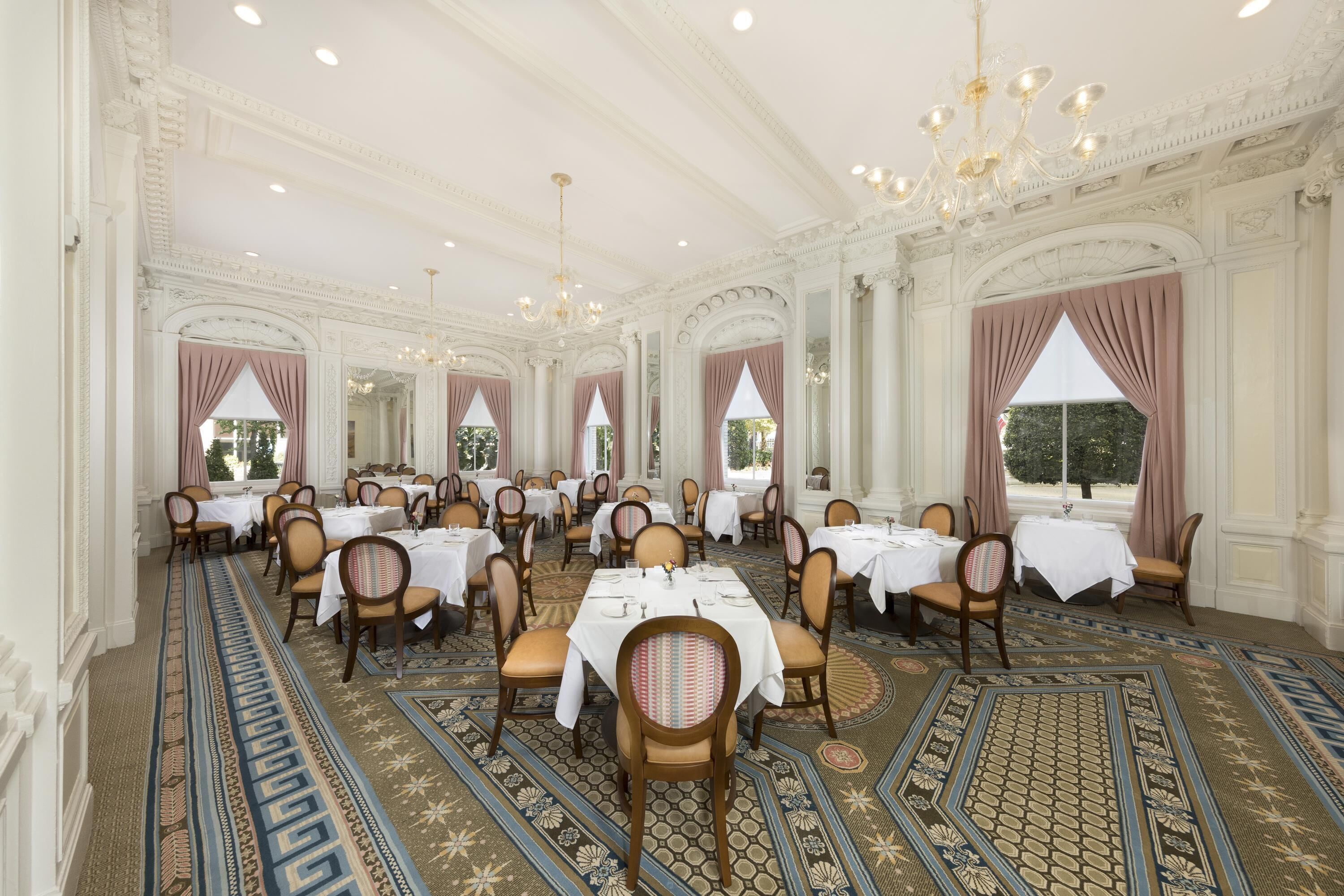 Un salón comedor imponente con columnas decorativas, candelabros y alfombra estampada. Las mesas están cubiertas con manteles blancos y sillas dispuestas.