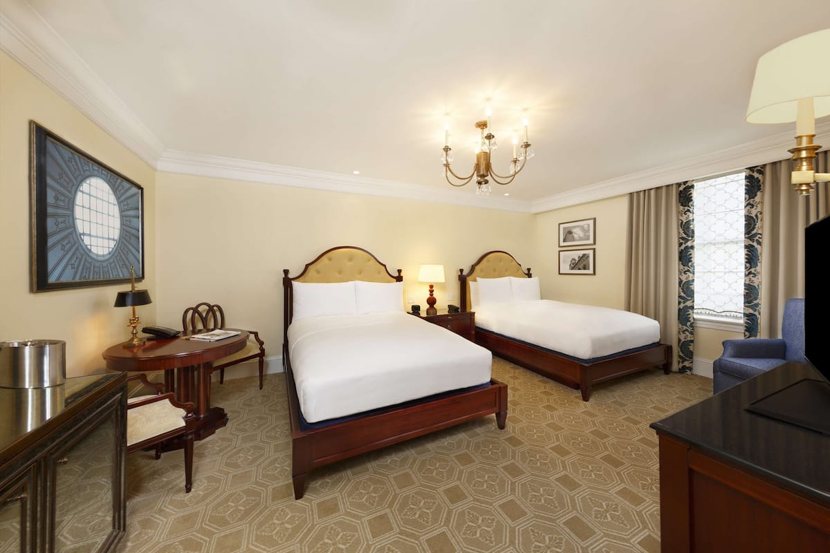 A hotel room with two beds, a desk, a chair, a TV, and a chandelier.