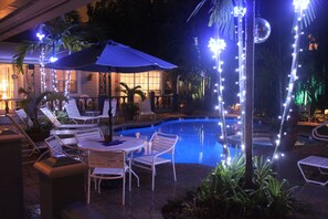 A poolside area with lounge chairs, a table with an umbrella, and string lights. - Coral Reef Guesthouse (Fort Lauderdale)