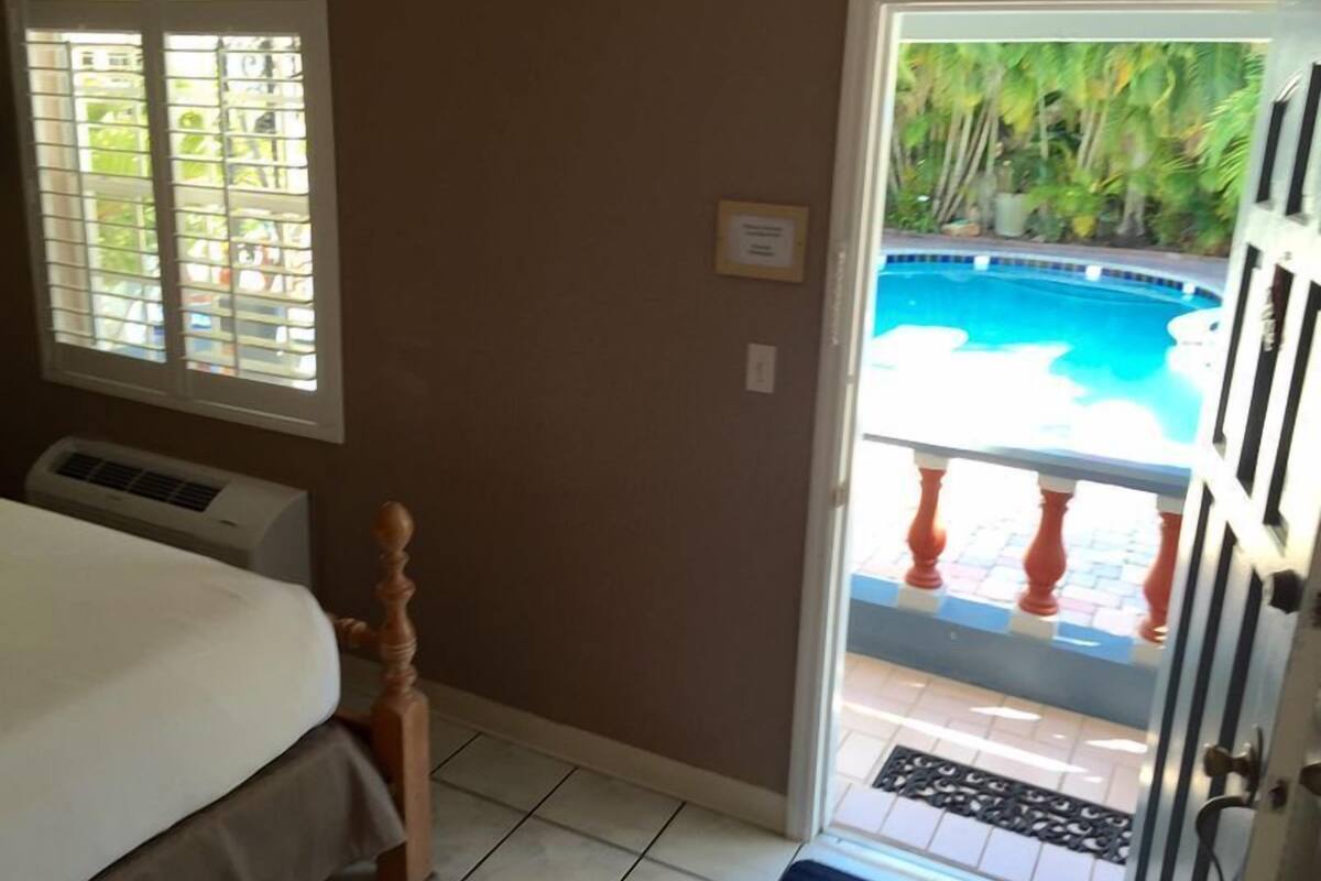 Une chambre avec un lit, une fenêtre avec des stores, un appareil de climatisation et une porte donnant sur un balcon avec vue sur la piscine.