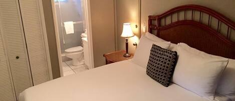 A hotel room with a bed, a nightstand with a lamp, a wooden headboard, and a view of the bathroom.
