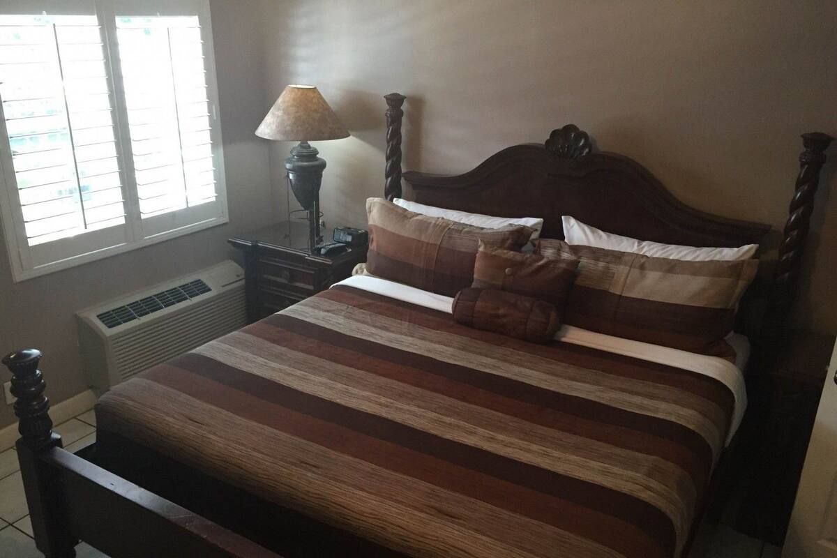 Une chambre avec un lit en bois, une table de chevet avec une lampe, une fenêtre avec des stores et un radiateur fixé au mur.